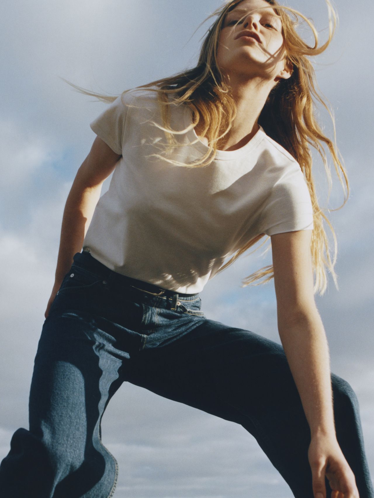 A person with long hair poses outdoors, wearing a white T-shirt and jeans, with a dynamic upward angle and a cloudy sky background.