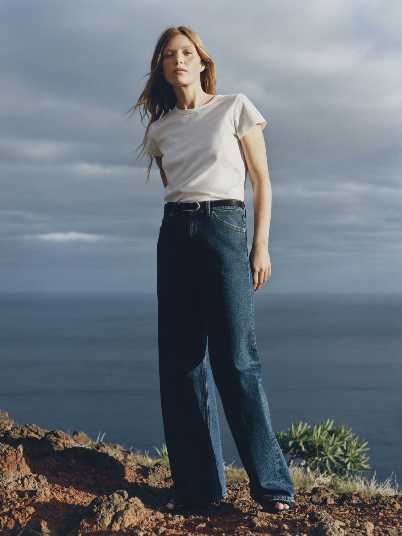 A person stands on a rocky cliff wearing a white T-shirt and dark jeans, with the ocean and cloudy sky in the background.