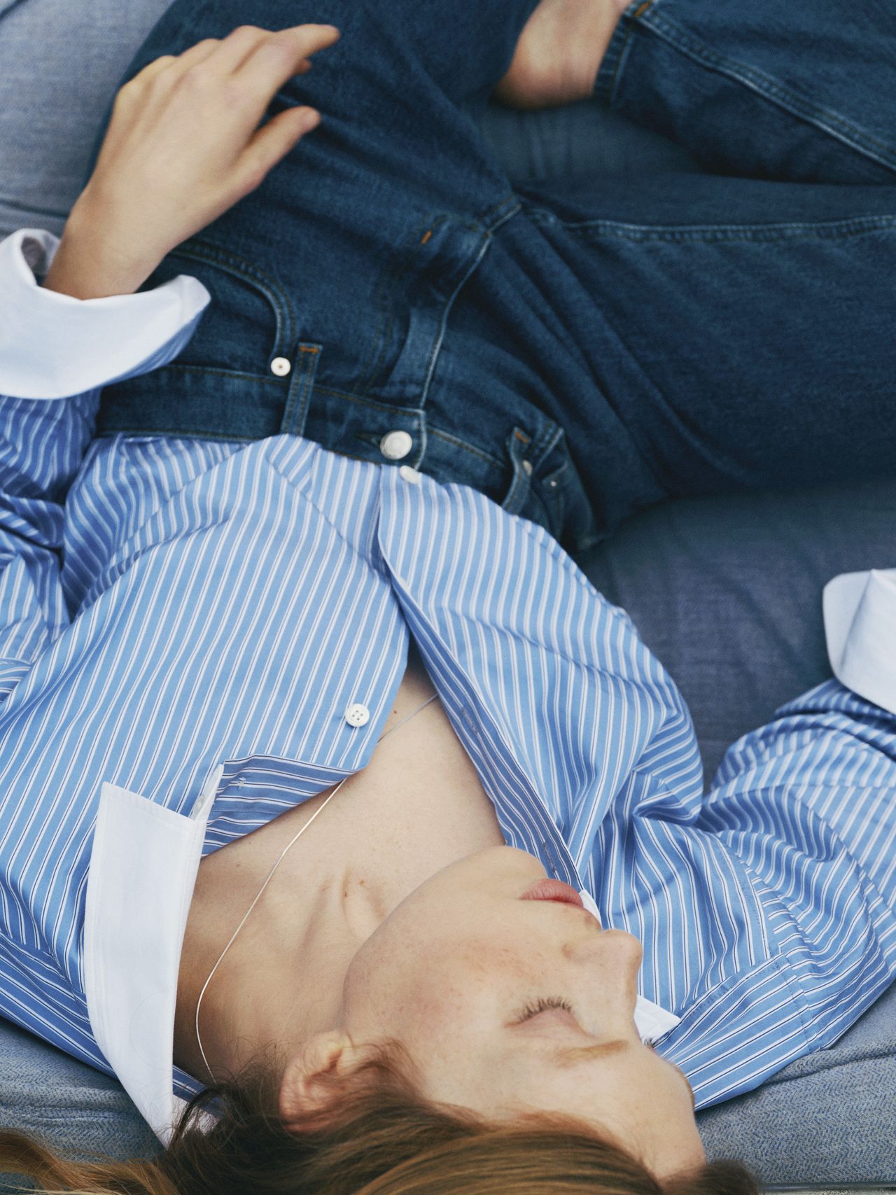 Person reclined on a sofa, wearing a blue striped shirt and jeans, with eyes closed and hair partially covering their face.