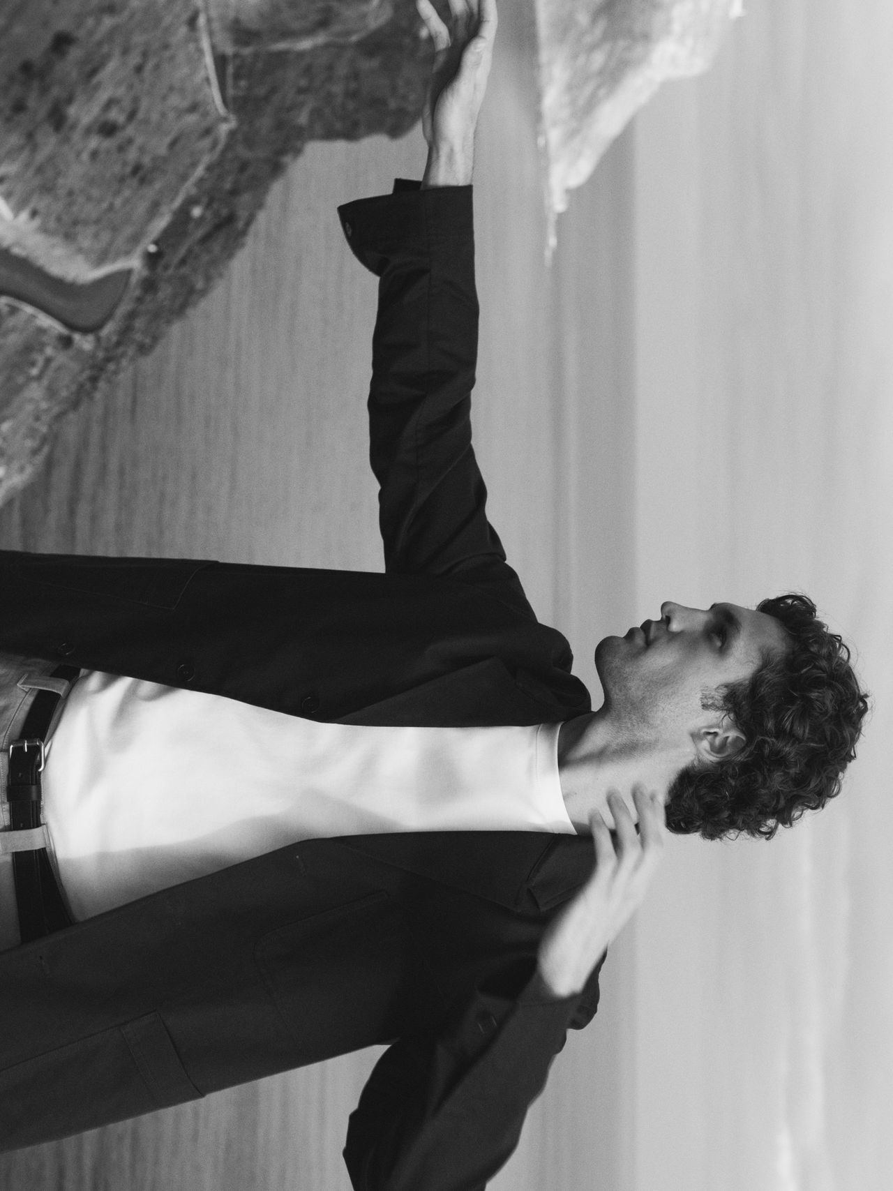 Man in a dark blazer gesturing by a rock formation near a body of water, in a black and white image.