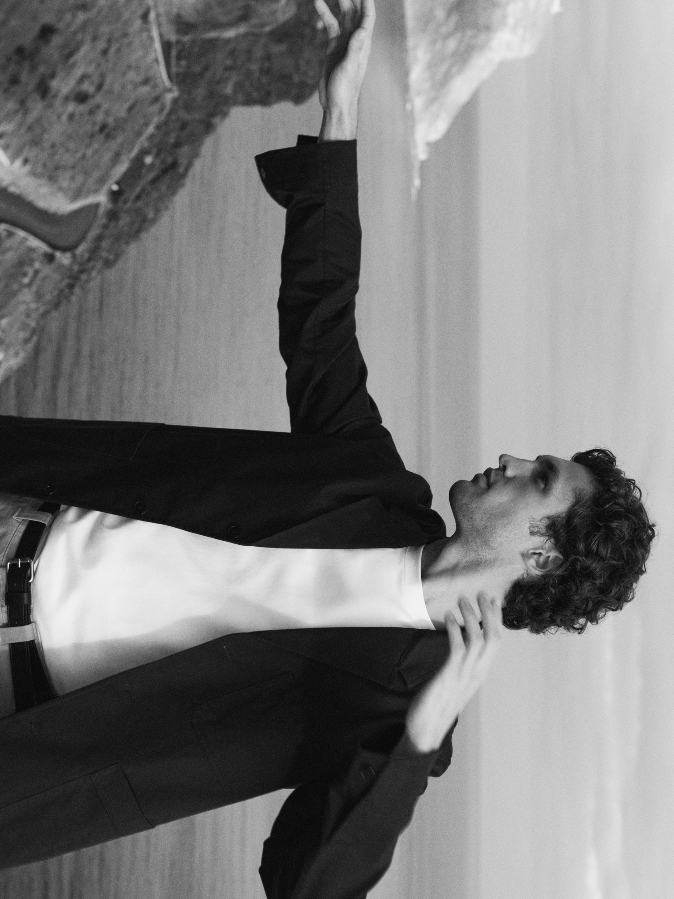 Man in a dark blazer gesturing by a rock formation near a body of water, in a black and white image.