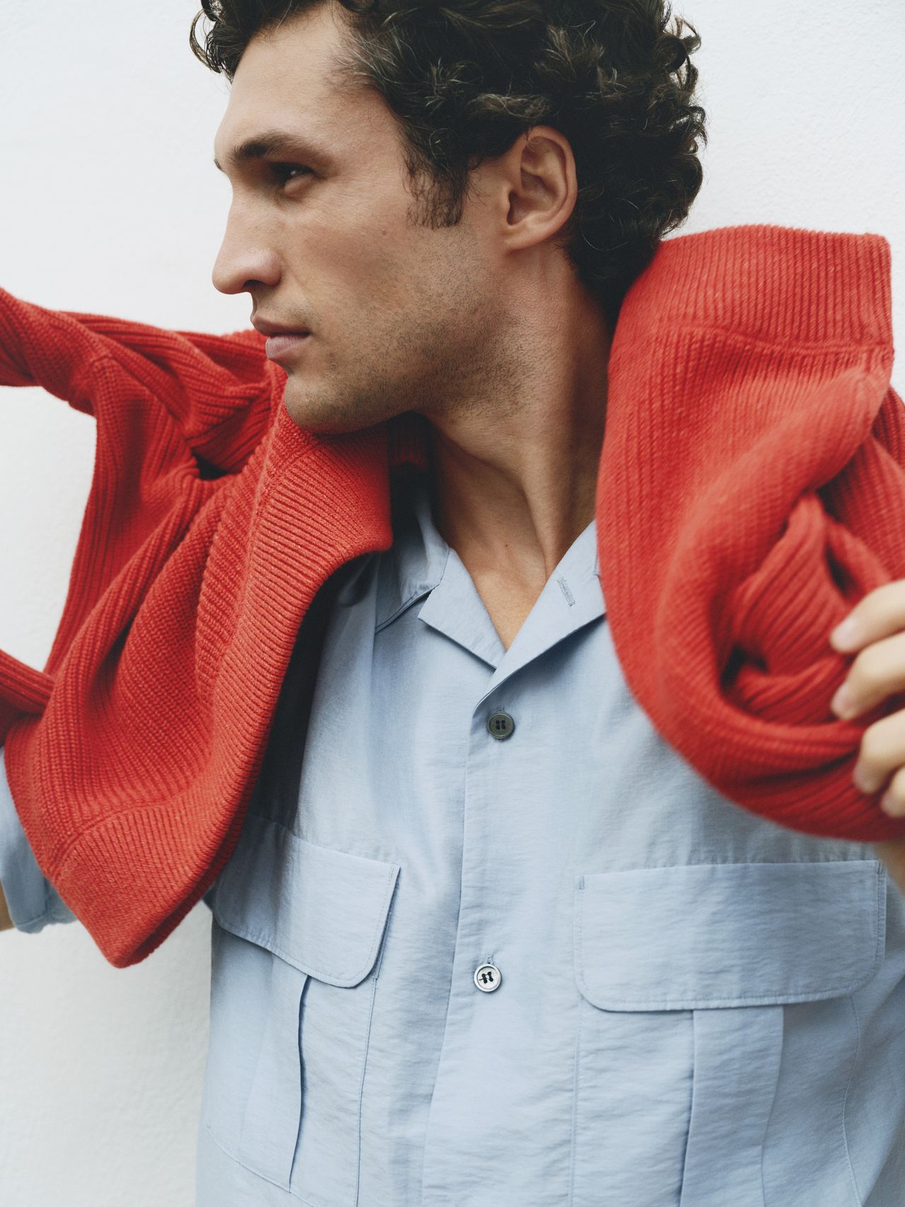 A man in a blue shirt drapes a red sweater over his shoulders, looking to the side against a plain white background.