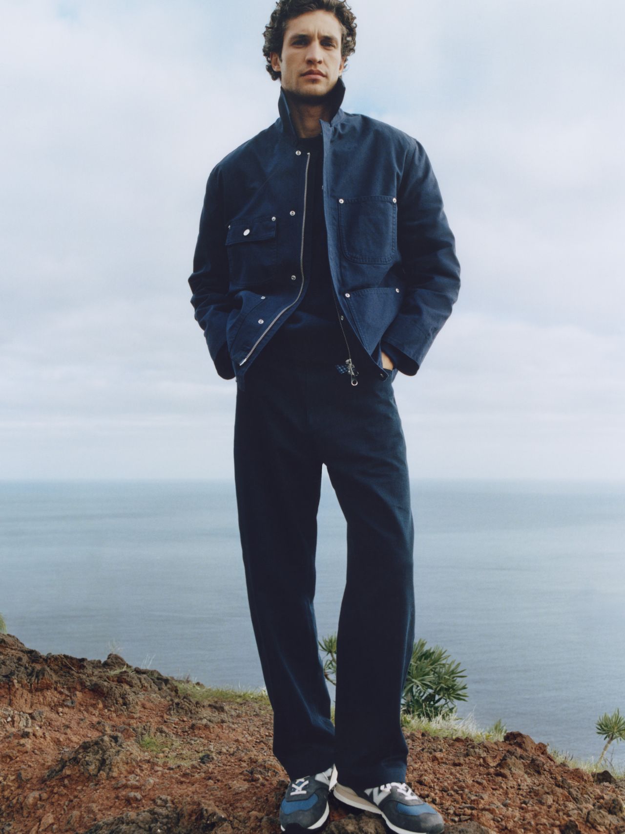 Person in a blue jacket and pants stands on a rocky terrain with the ocean and cloudy sky in the background.