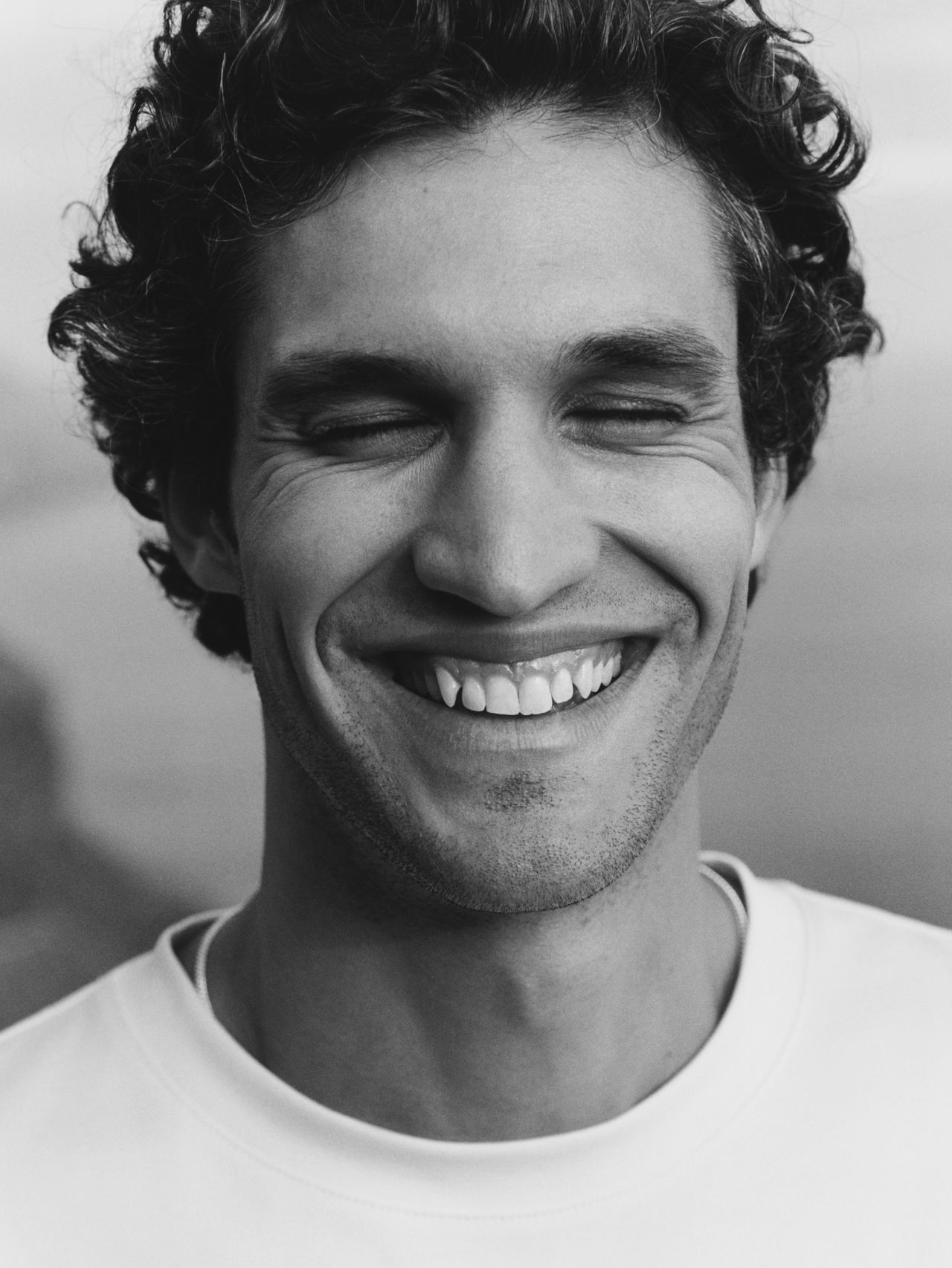 Black and white photo of a smiling man with curly hair, eyes closed, wearing a light-colored shirt.