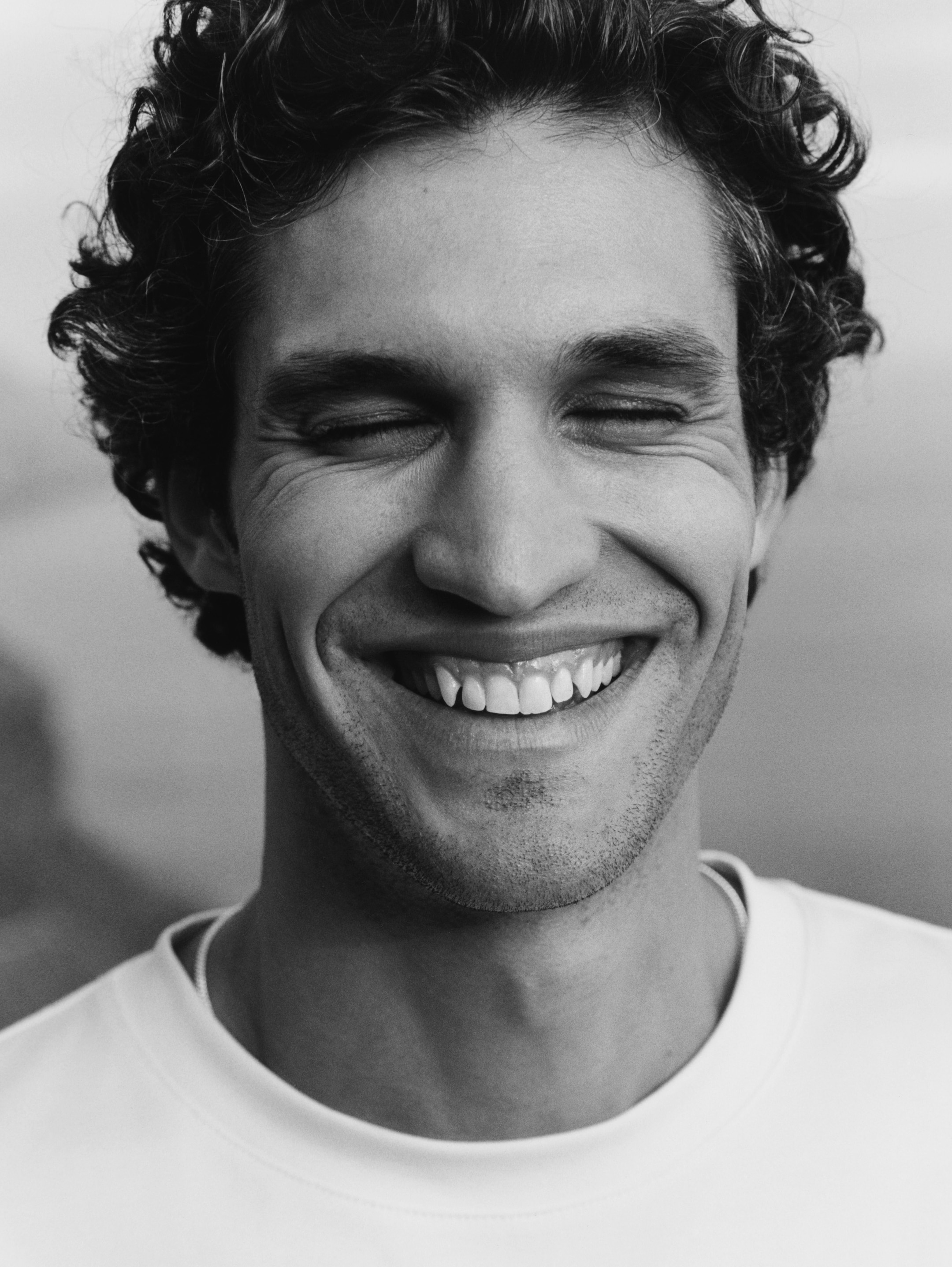 Black and white photo of a smiling man with curly hair, eyes closed, wearing a light-colored shirt.