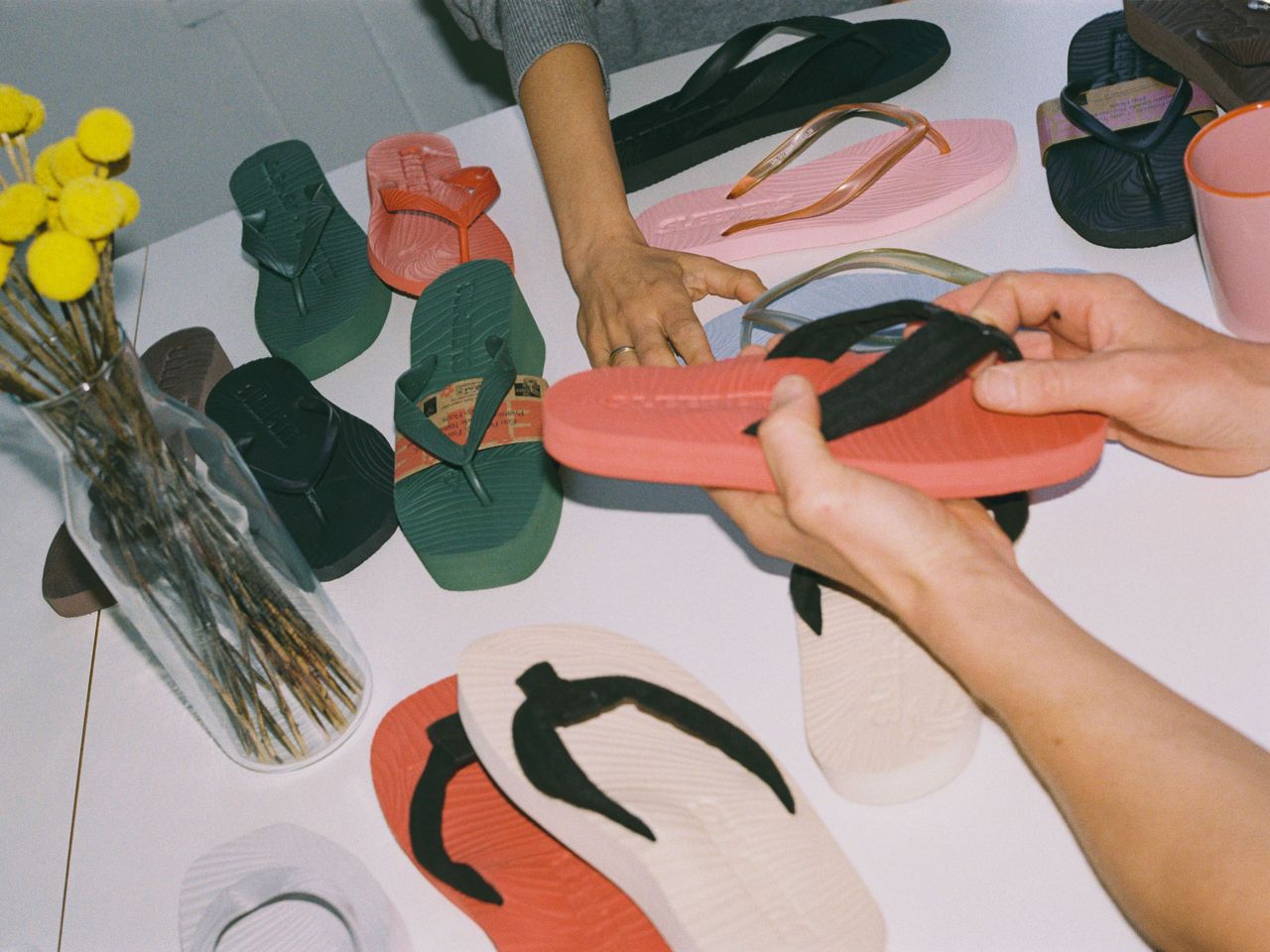 Hands handling colorful flip-flops on a table with more footwear and a vase of yellow flowers in the background.