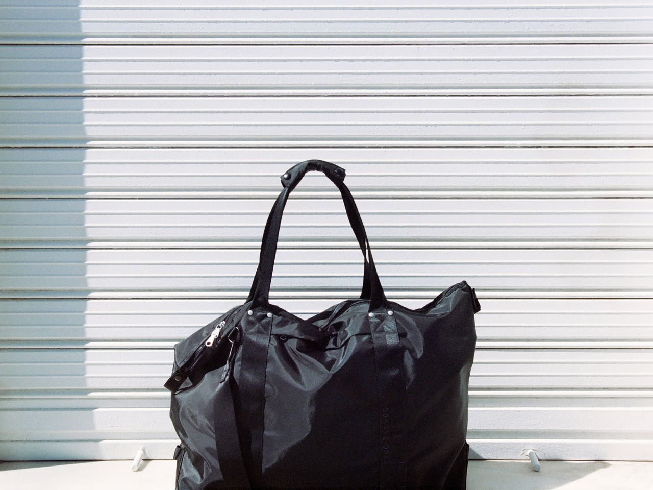 A large black duffel bag with handles sits in front of a white, corrugated metal background, casting a shadow on the surface.