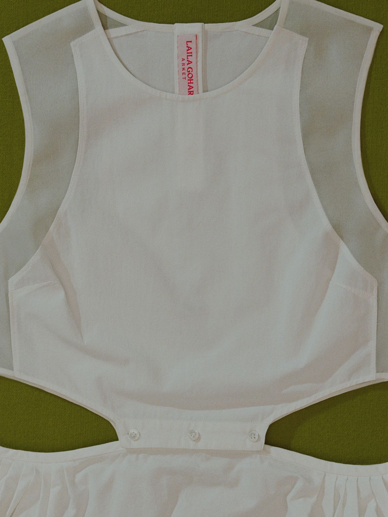 White sleeveless dress with sheer shoulder panels and cutout waist on a green surface, featuring a red label inside the neck area.