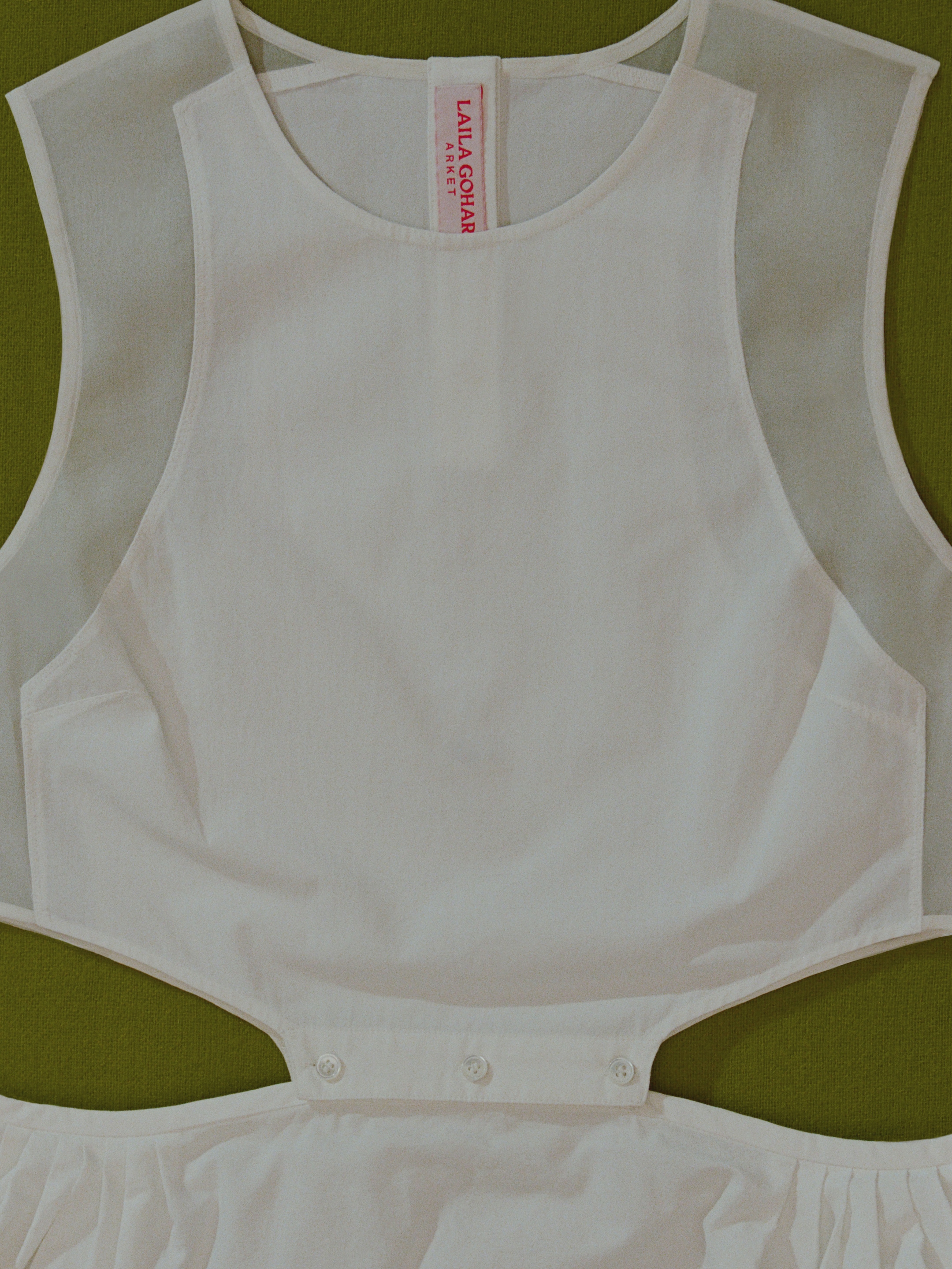 White sleeveless dress with sheer shoulder panels and cutout waist on a green surface, featuring a red label inside the neck area.