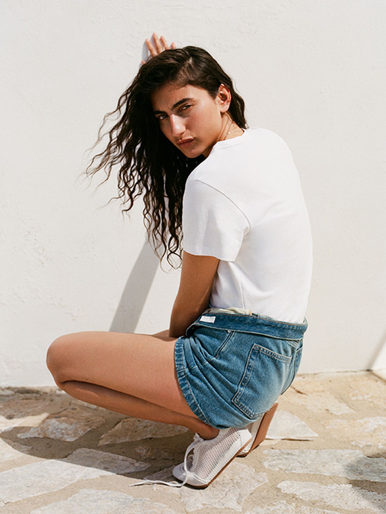 A woman with long curly hair crouches against a white wall, wearing a white T-shirt, denim shorts, and white shoes, looking over her shoulder.