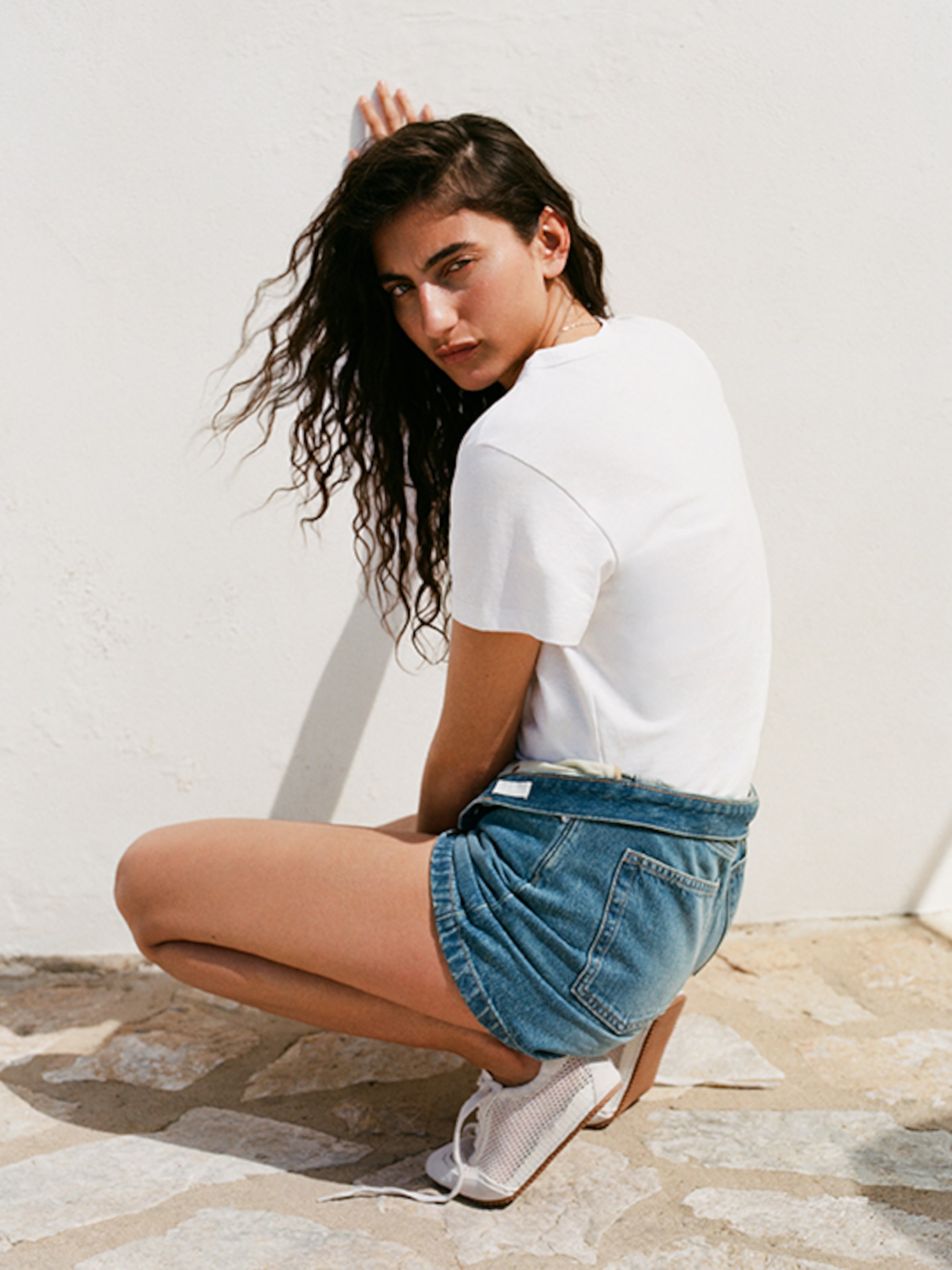 A woman with long curly hair crouches against a white wall, wearing a white T-shirt, denim shorts, and white shoes, looking over her shoulder.
