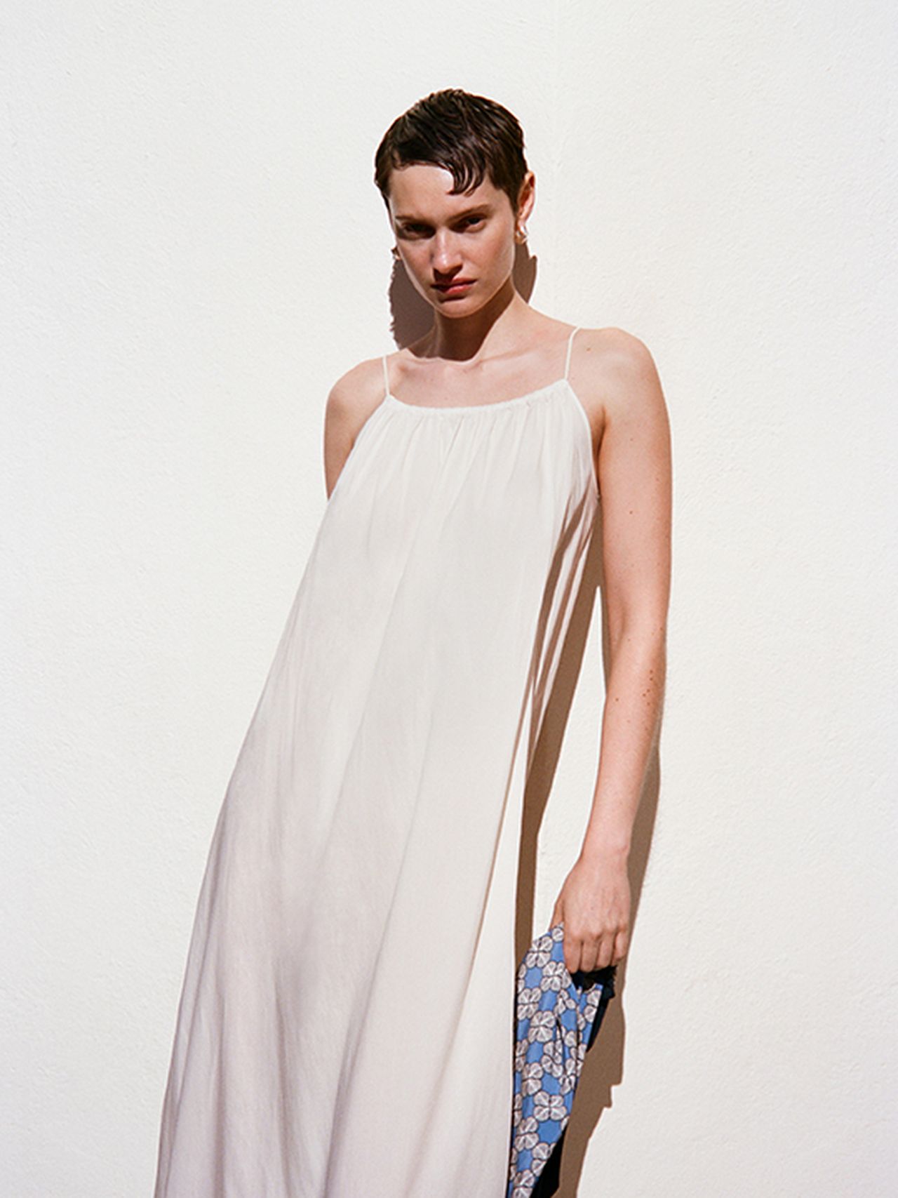 Woman in a white spaghetti strap dress stands against a white wall, holding a blue patterned scarf, with short hair styled neatly.