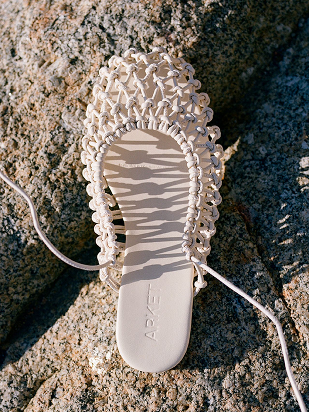 Beige woven sandal on a textured rock surface, casting a shadow, with "ARKET" embossed on its footbed.