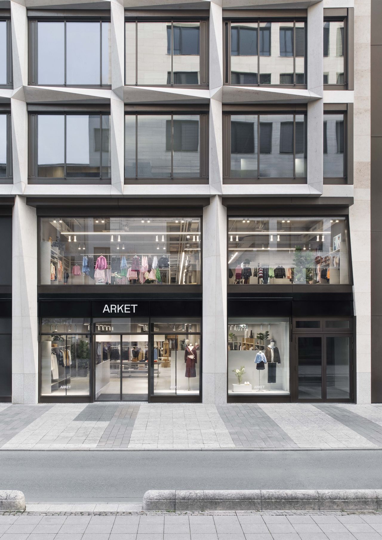 Street view of a modern ARKET store with large glass windows displaying clothing and accessories on two floors.