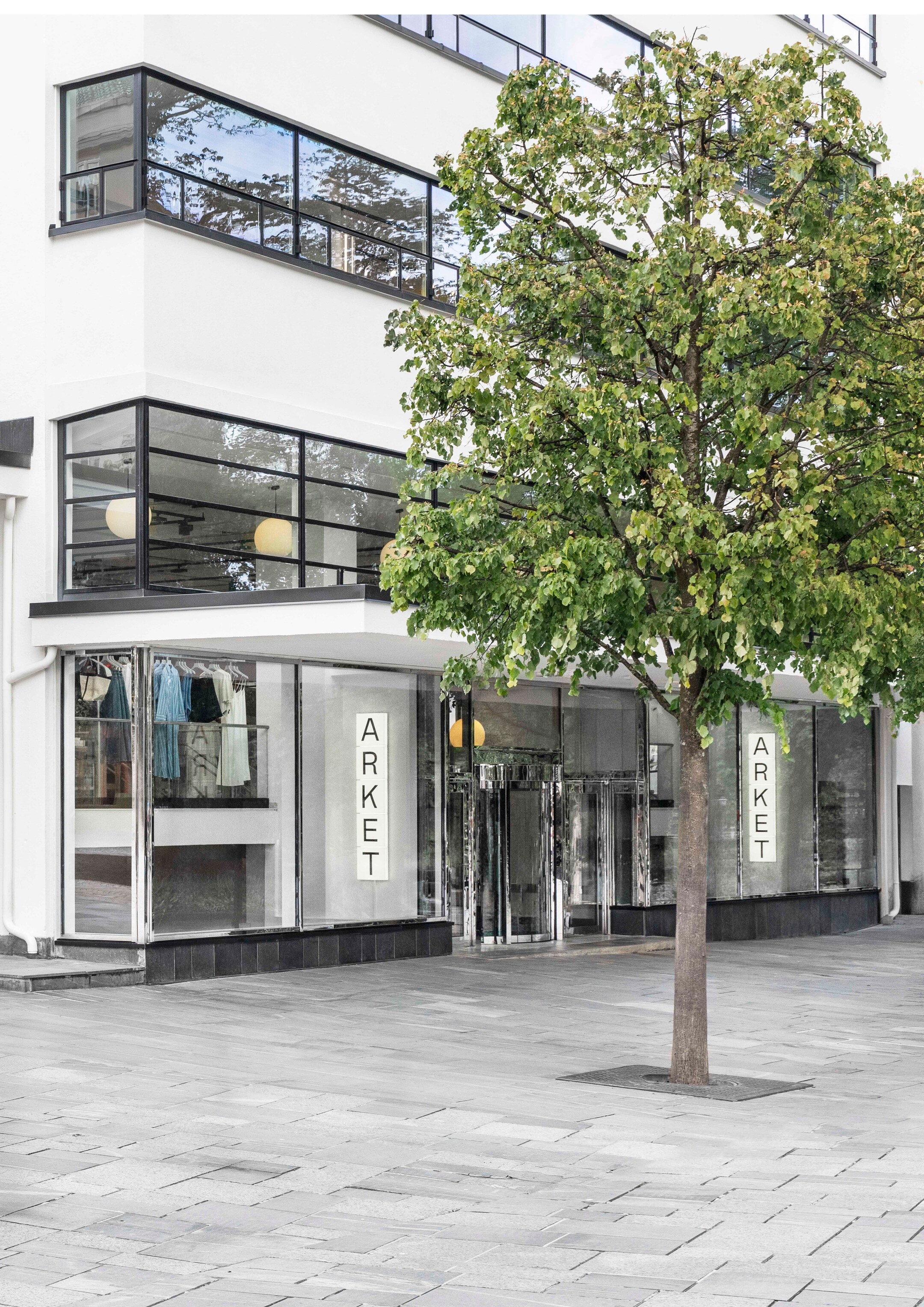 Modern storefront with large windows displaying clothing, labeled "ARKET." A leafy tree stands in front on a paved sidewalk.