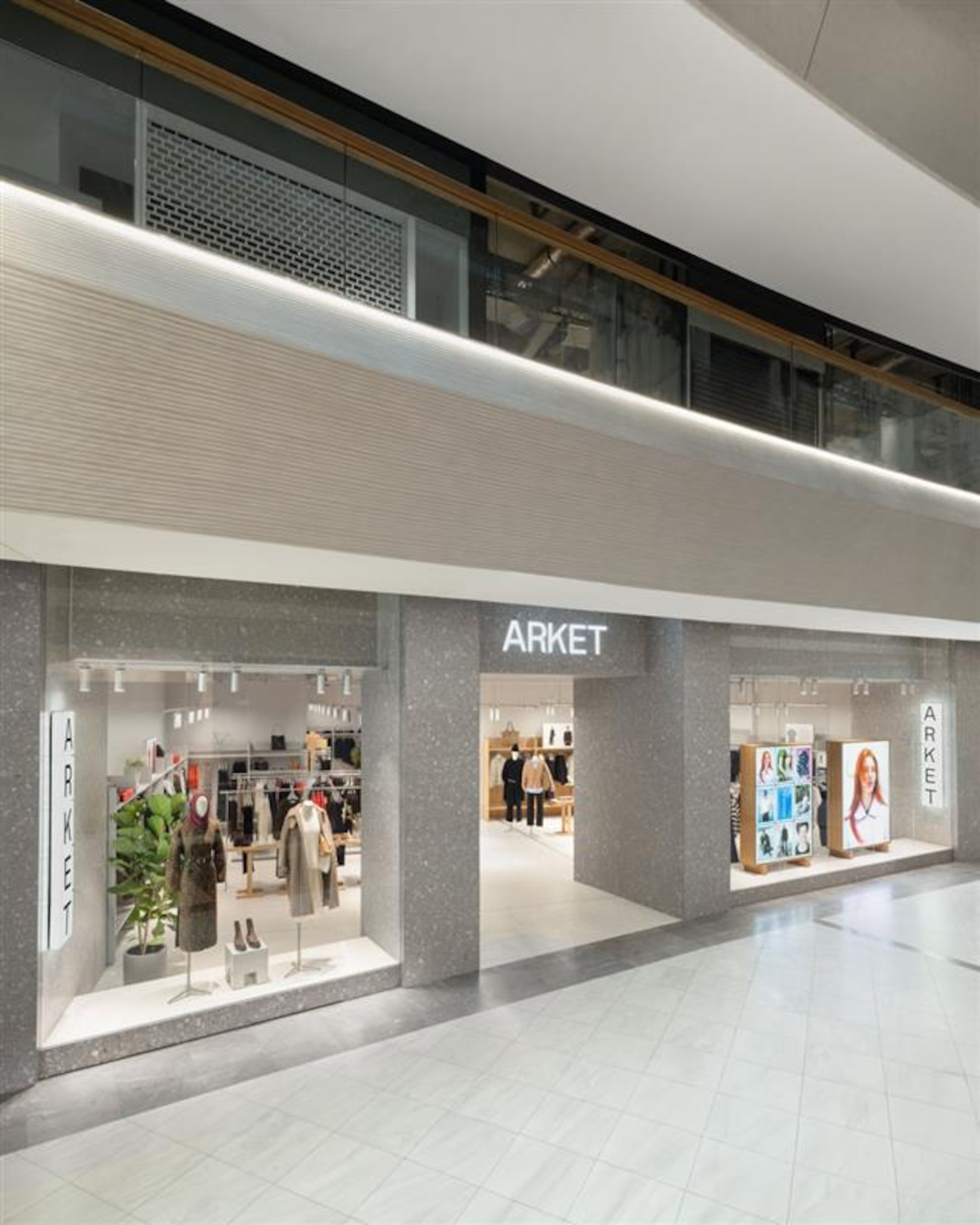 Storefront of ARKET in a mall, featuring mannequins, clothing displays, and digital screens showcasing fashion images. Modern and minimalist design.