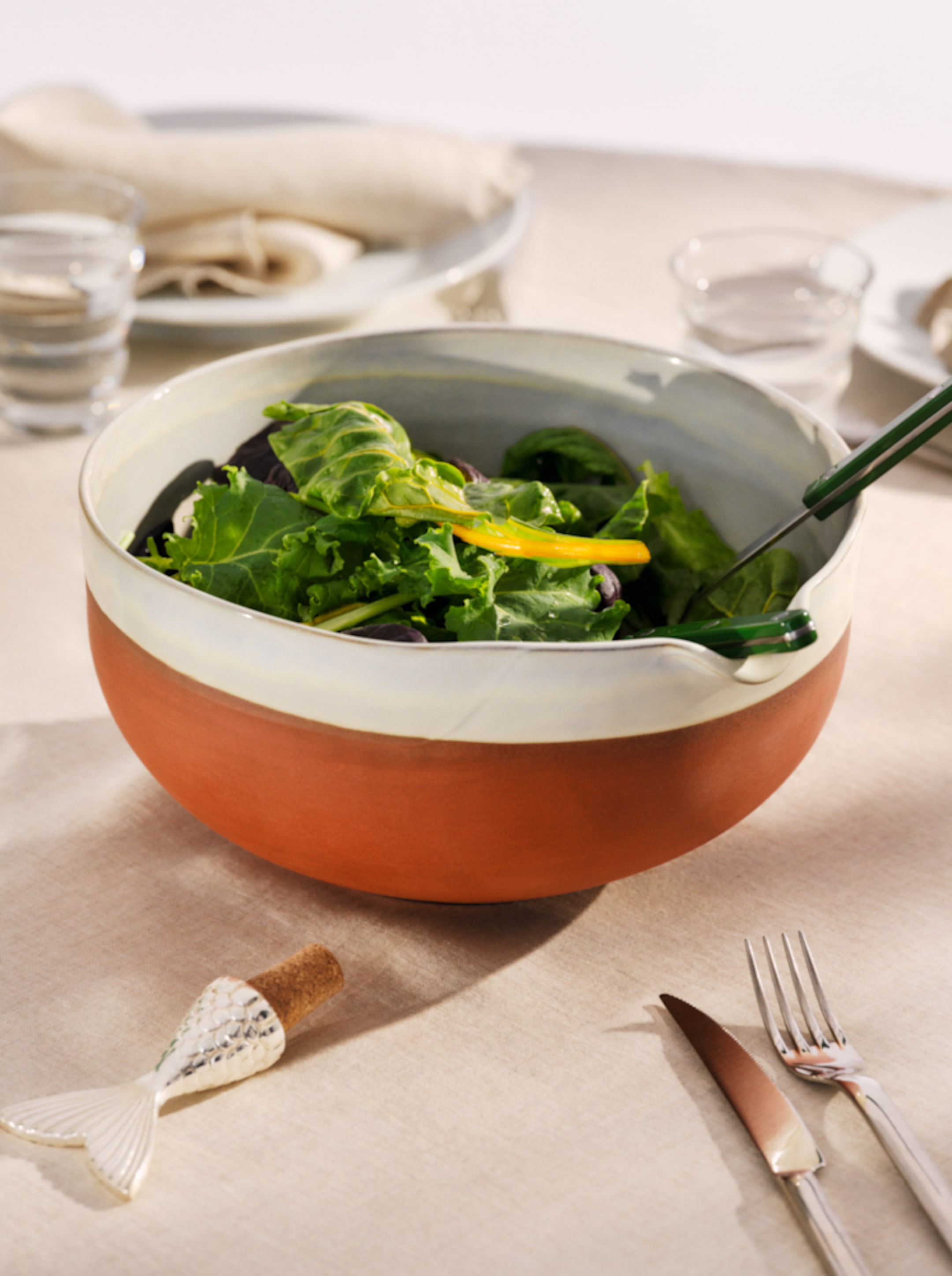 A ceramic bowl of fresh green salad with yellow bell pepper on a laid table with cutlery, a cork wine stopper, and glasses of water.