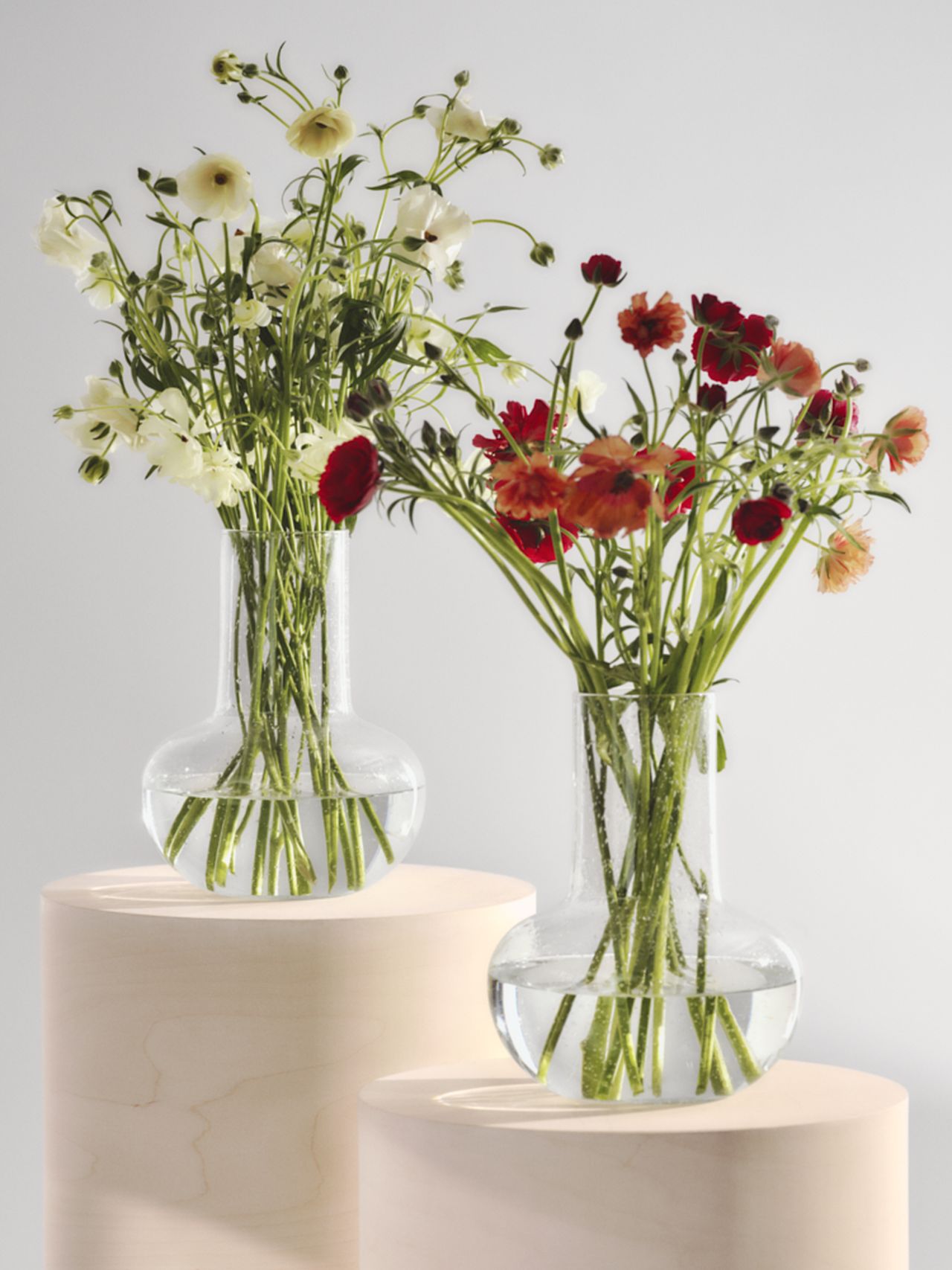 Two glass vases with red and white flowers on wooden pedestals against a light gray background.