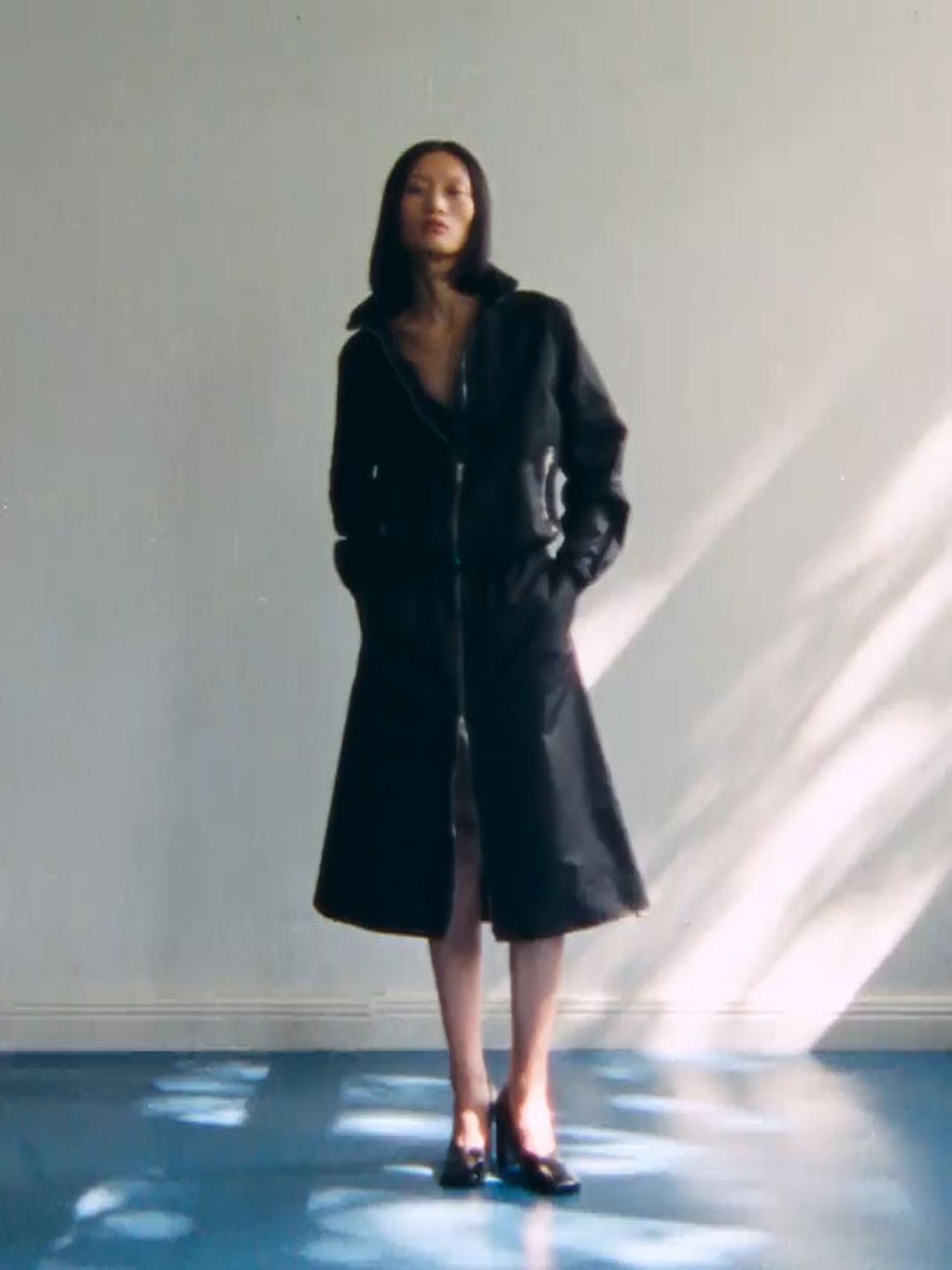 Person standing in a black coat and heels against a light blue wall, with sunlight creating patterns on the floor.