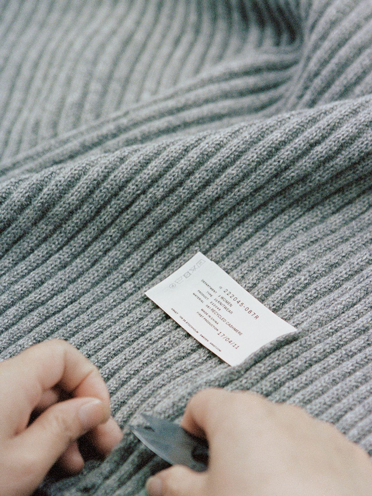 Hands cutting a tag from a gray ribbed fabric garment with a pair of scissors, focusing on the texture and label details.
