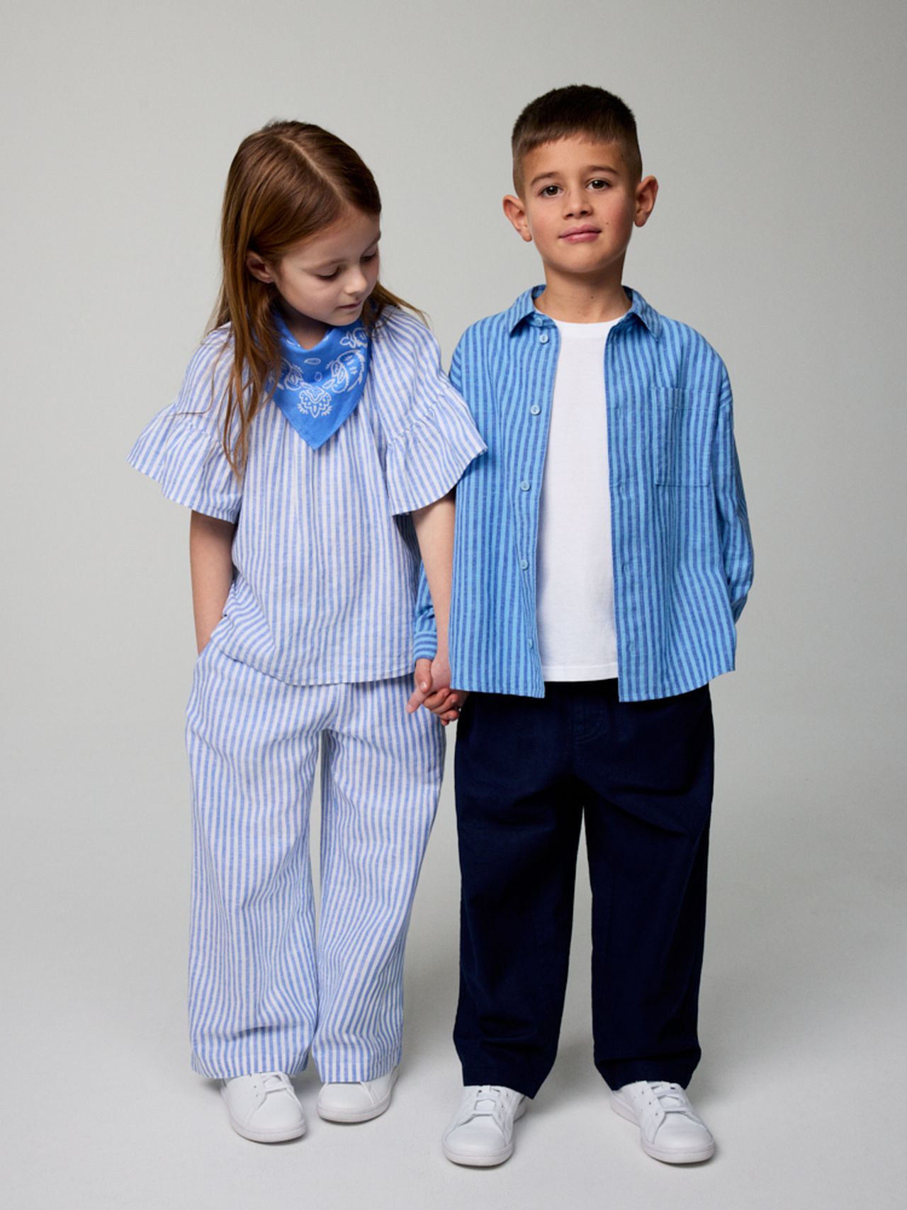 A young girl and boy in matching blue striped outfits and white shoes stand holding hands against a plain background.