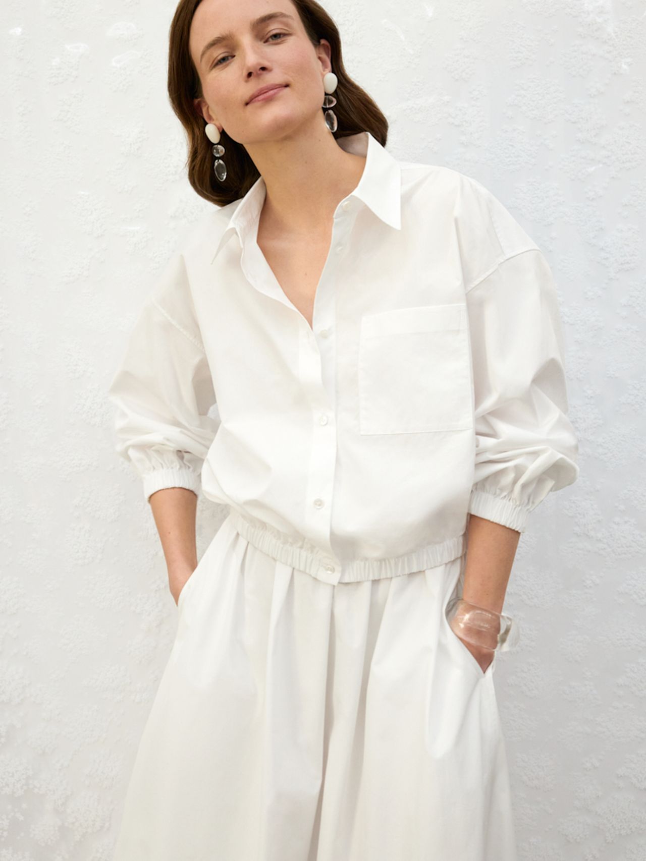 Person wearing a loose white button-up shirt and matching skirt, hands in pockets, with pearl earrings against a white textured background.