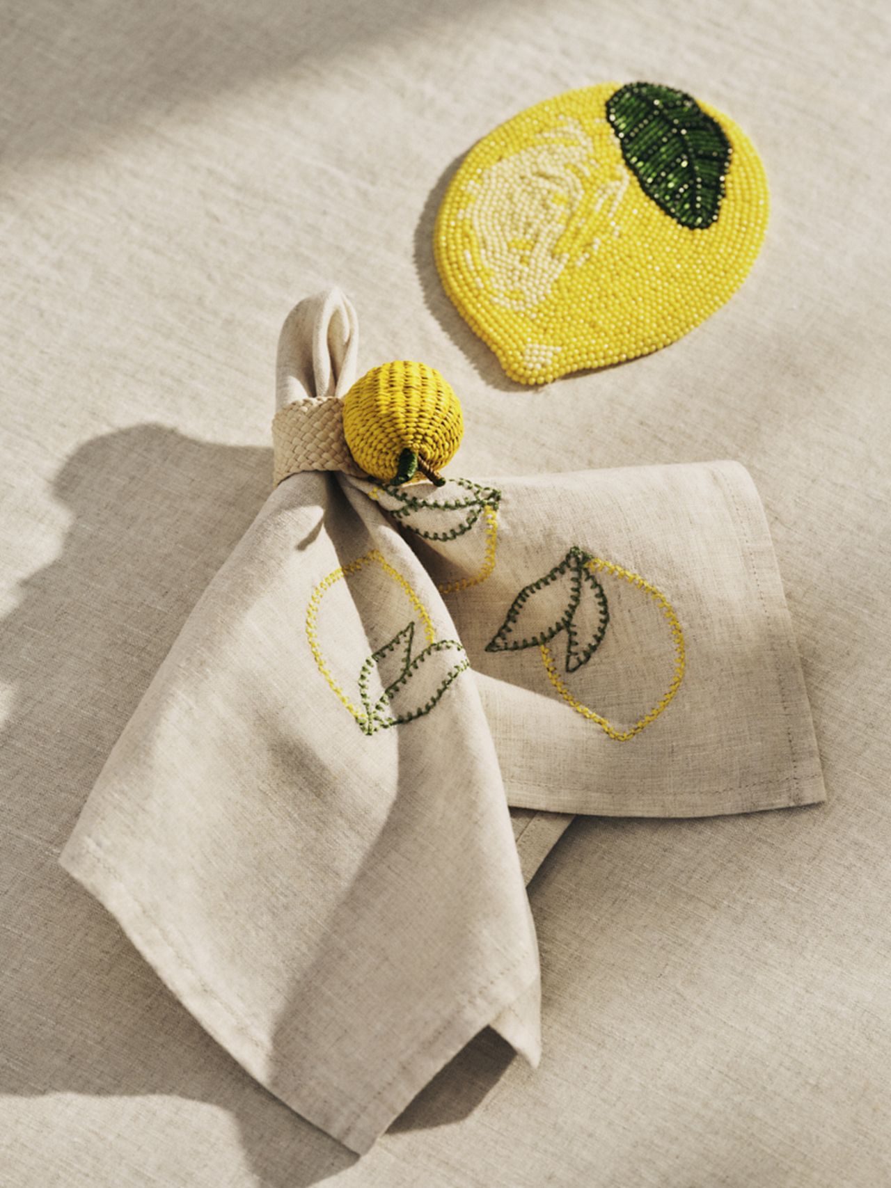Linen napkin with embroidered lemons and a woven lemon napkin ring beside a beaded lemon coaster on a beige tablecloth.