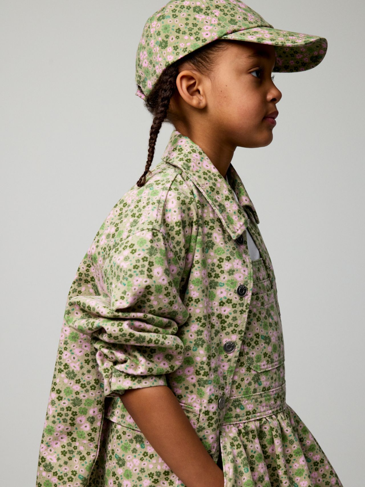 Young girl in profile wearing a green and pink floral outfit with a matching cap, looking to the left against a neutral background.