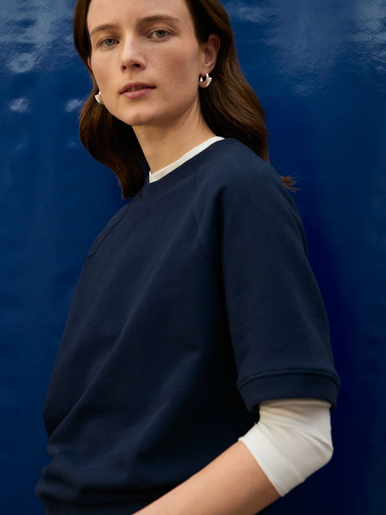 Person with brown hair, wearing a navy blue top and hoop earrings, stands against a glossy blue background.
