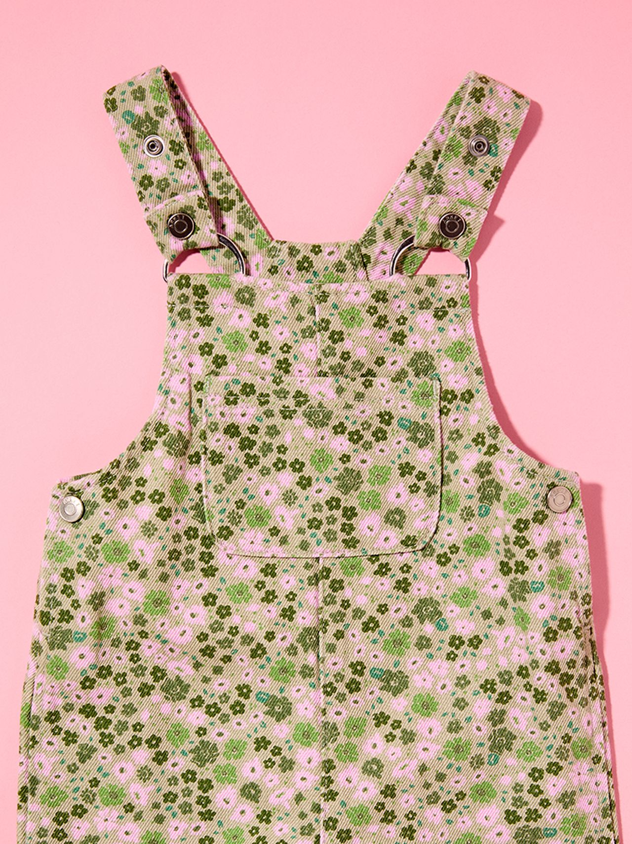 Green floral-patterned overalls with pink flowers, featuring metal button closures and a front pocket, set against a pink background.