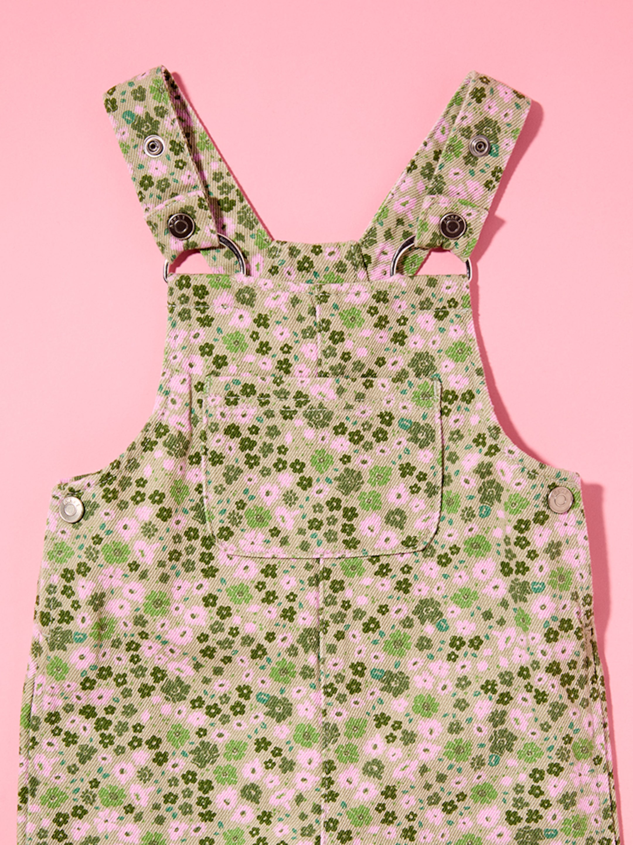 Green floral-patterned overalls with pink flowers, featuring metal button closures and a front pocket, set against a pink background.