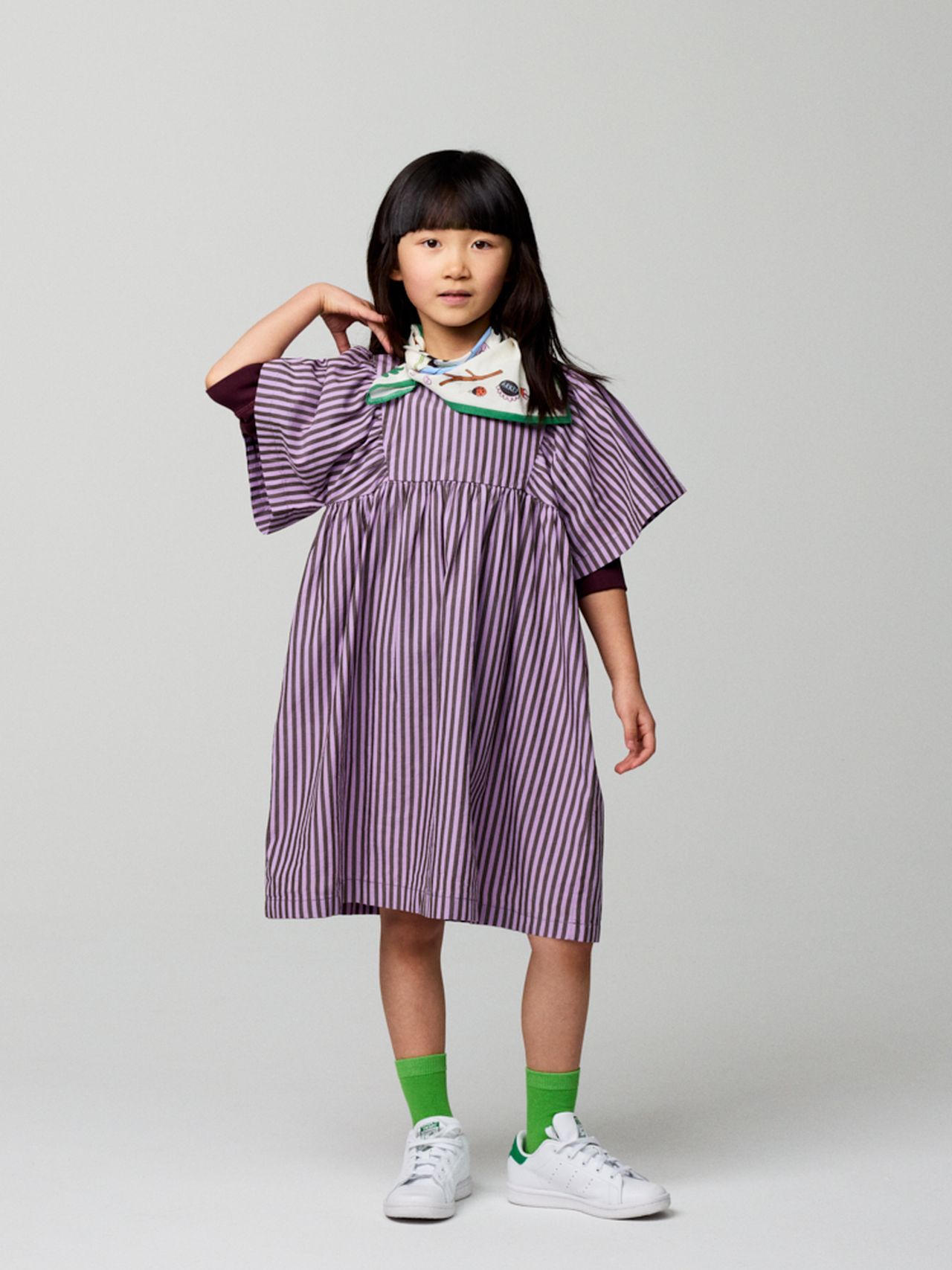 Young girl in a purple striped dress and green socks stands on a gray background, adjusting her shoulder. She wears white sneakers and a colorful scarf.