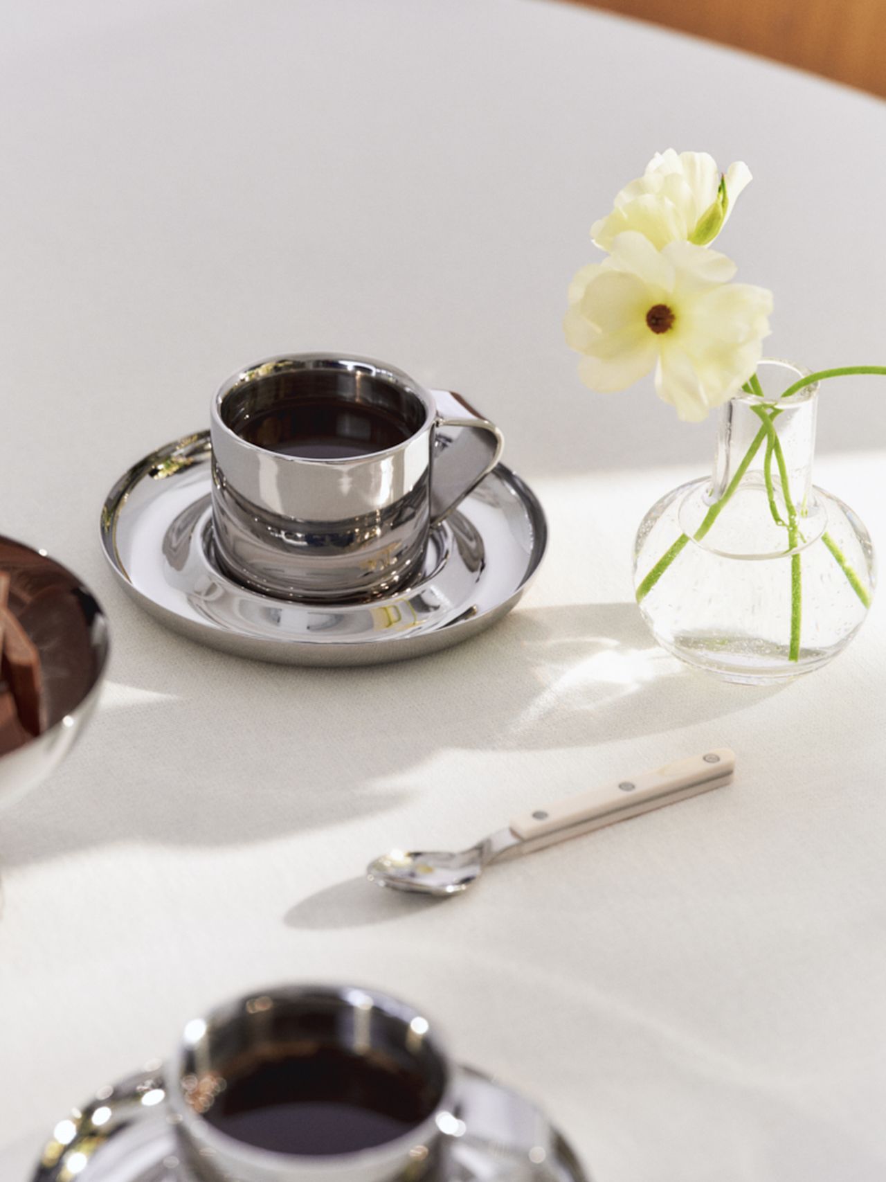 A metallic cup of coffee on a saucer sits beside a glass vase with a white flower and a small spoon on a white tablecloth.