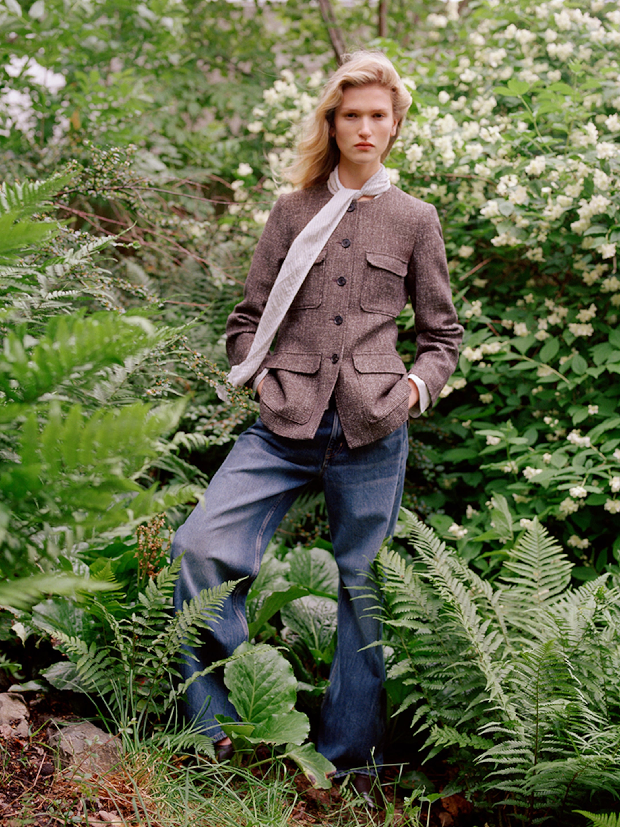 A person in a brown jacket and jeans stands among lush green plants and white flowers, with a serene expression, surrounded by vibrant foliage.