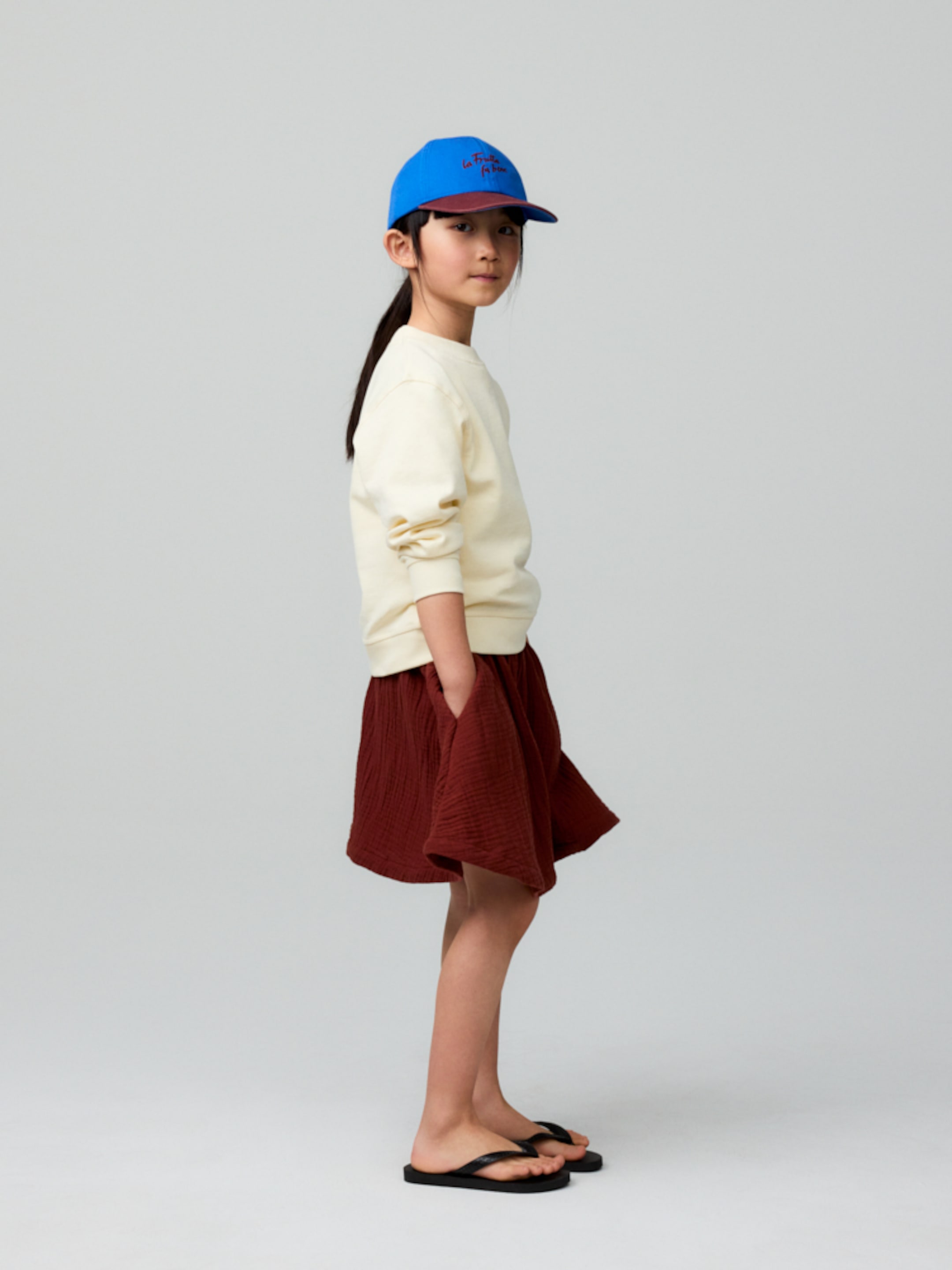 Young girl in a blue cap, cream sweater, and red skirt stands sideways in flip-flops. Minimalist gray background.