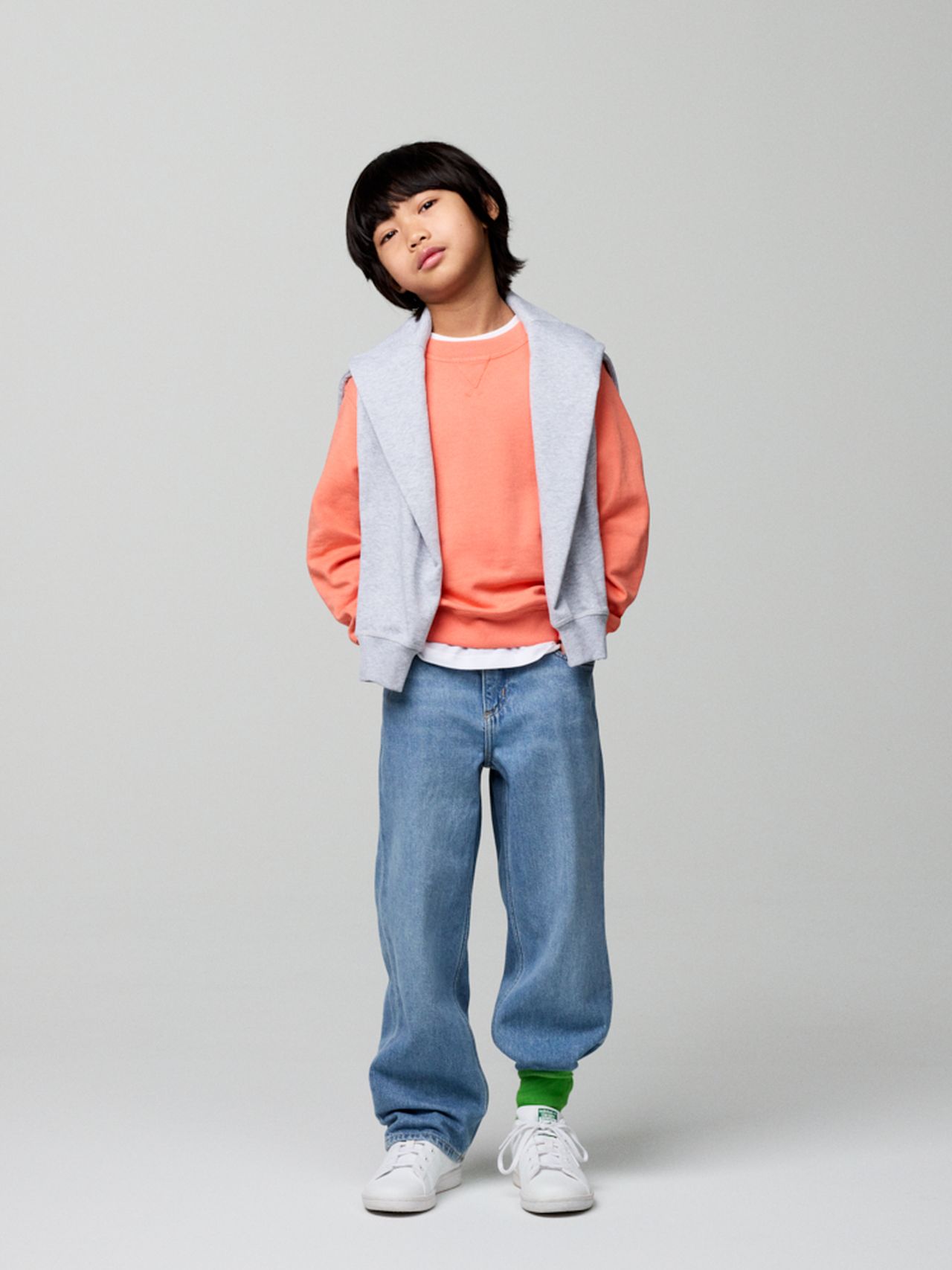 Child in a coral sweater and blue jeans, with a gray jacket draped over shoulders, stands confidently against a gray background.