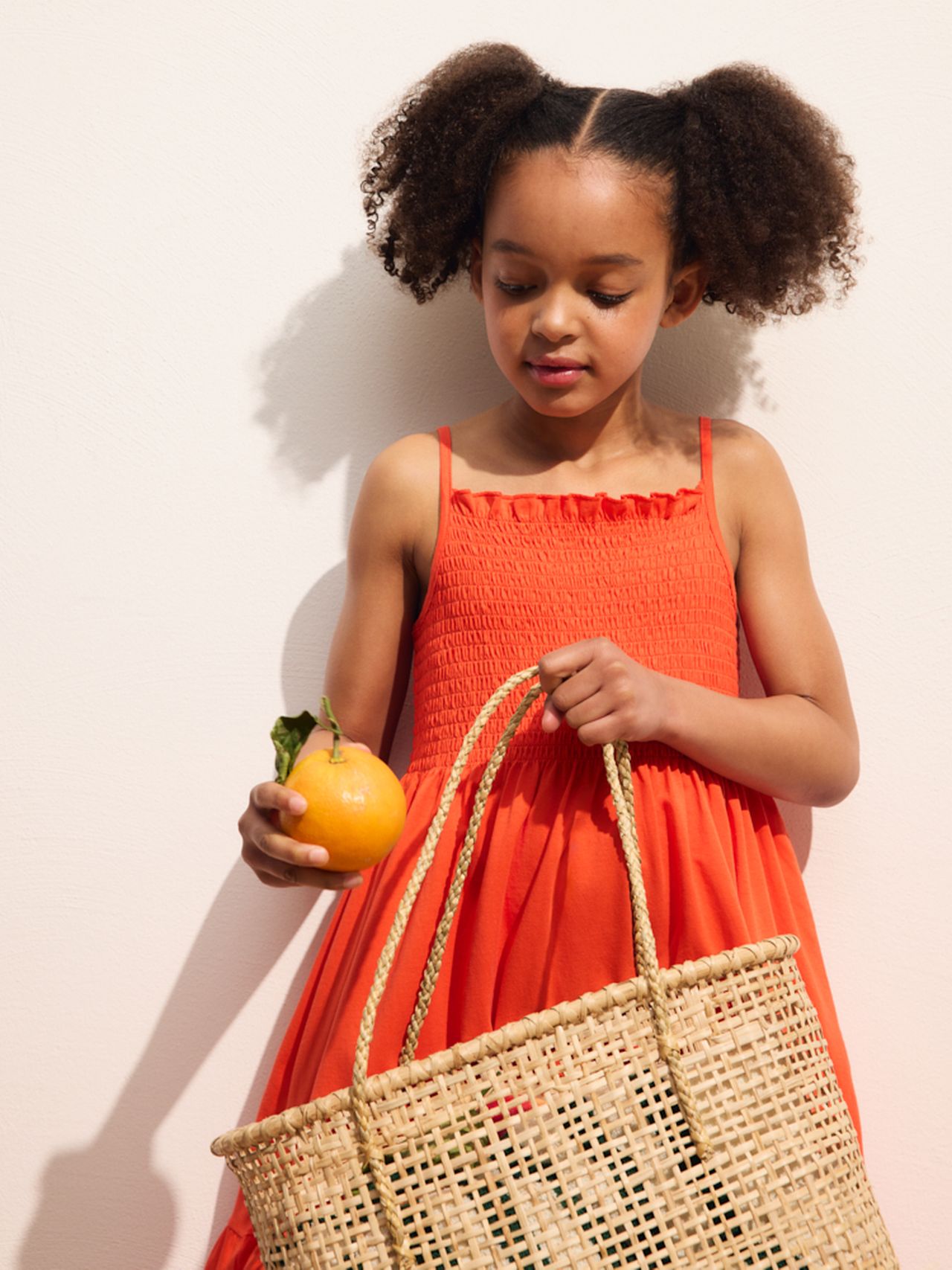Young girl in an orange dress holds an orange and a woven basket, standing against a light background.