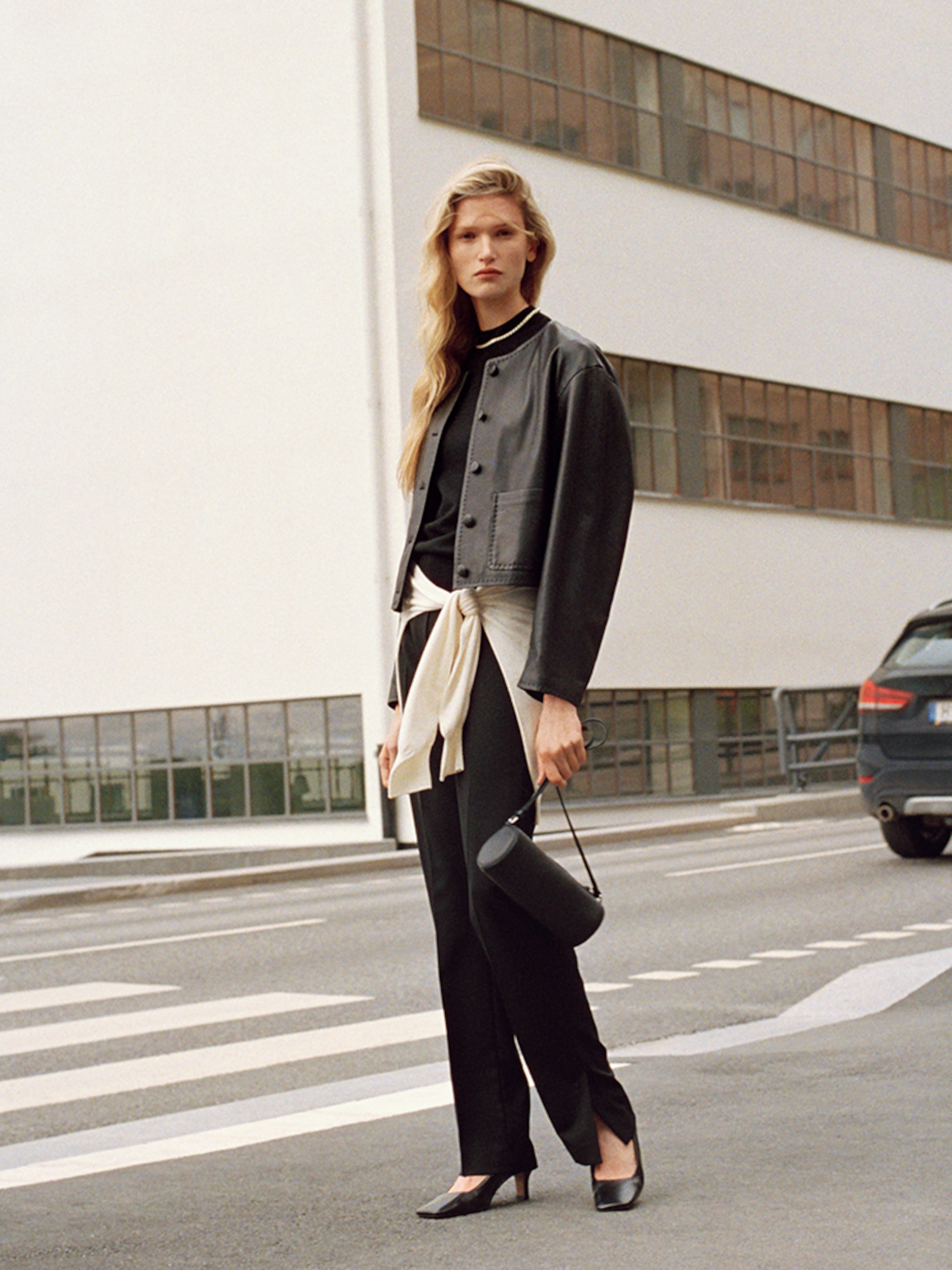 A woman stands confidently on a street, wearing a black outfit with a leather jacket and holding a small black bag. A modern building is in the background.