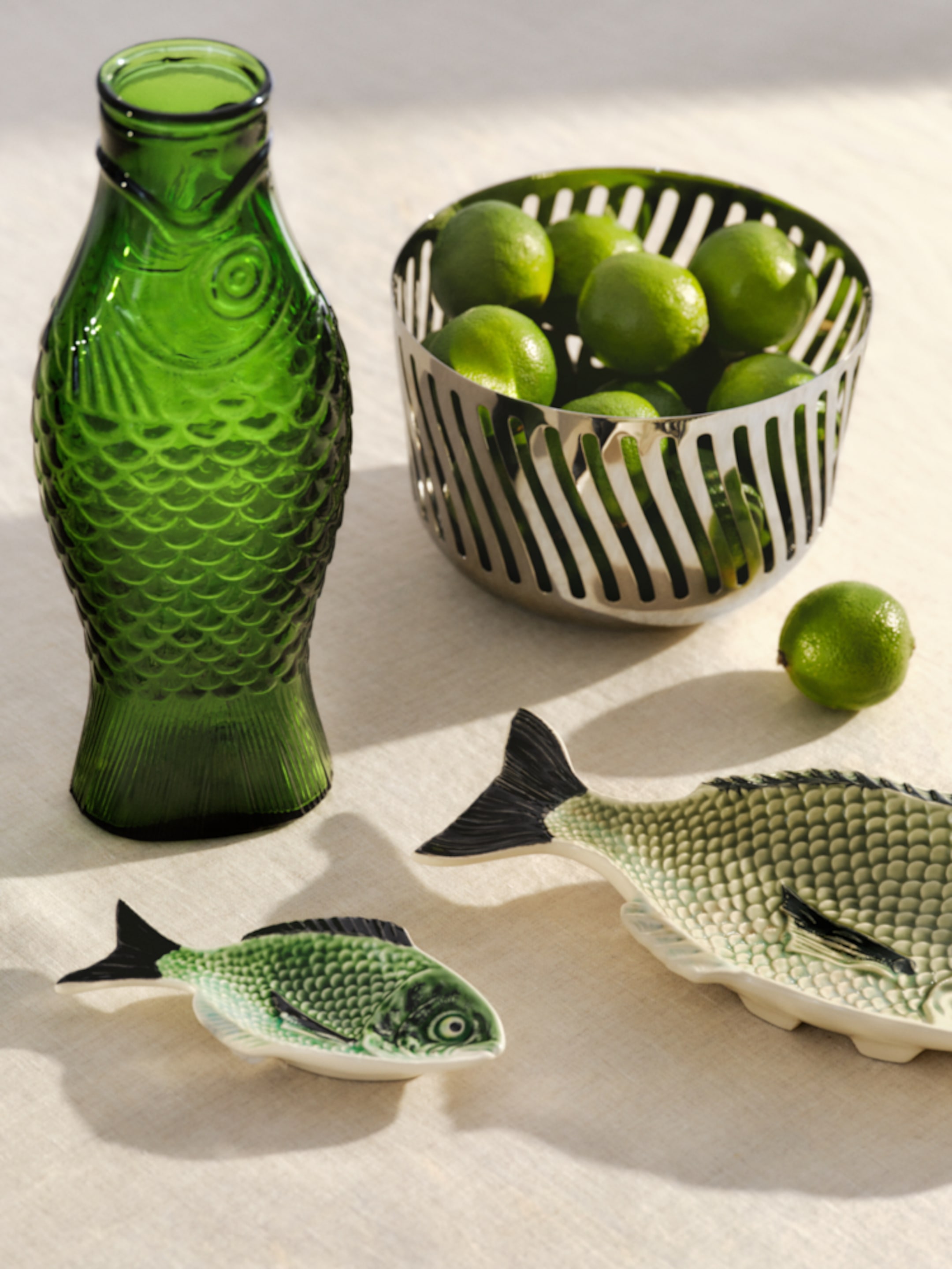 Green glass bottle, metal bowl with limes, and fish-shaped ceramic plates on a light tablecloth.