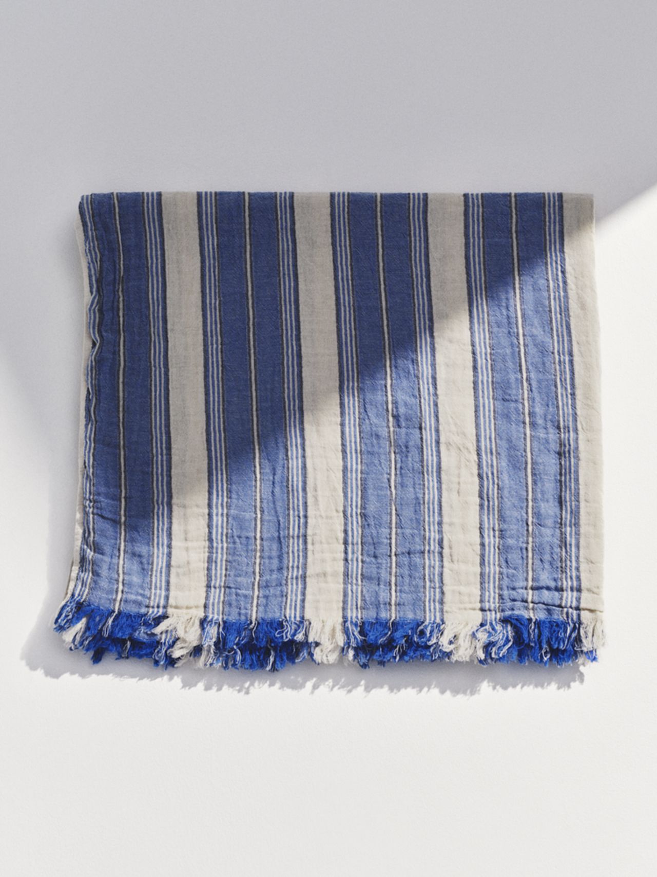 Folded blue and white striped fabric with fringed edges, laying flat on a white surface.