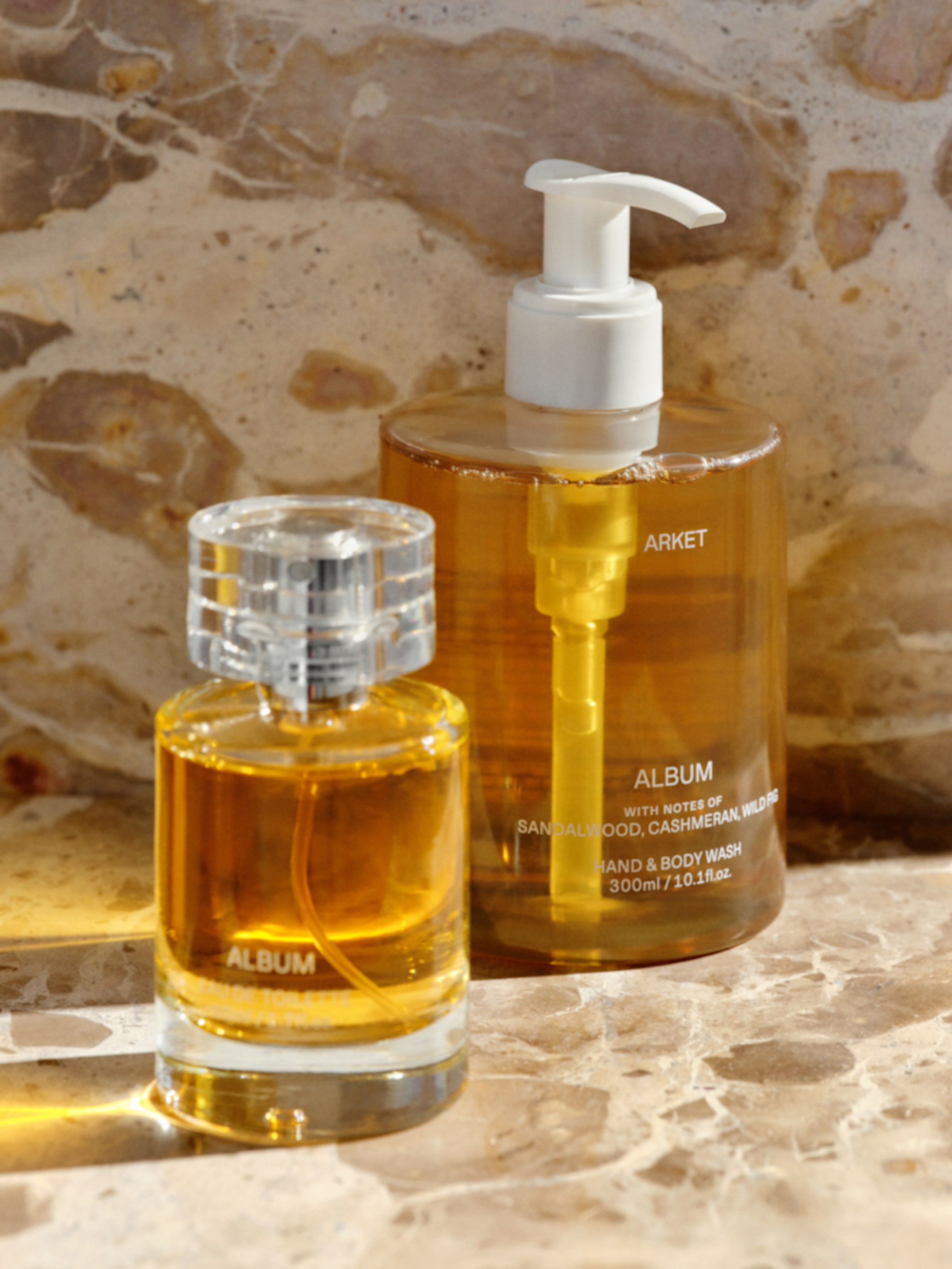 Two amber-colored bottles, one perfume and one hand and body wash, against a stone background. Labels read "Album" with notes of sandalwood.