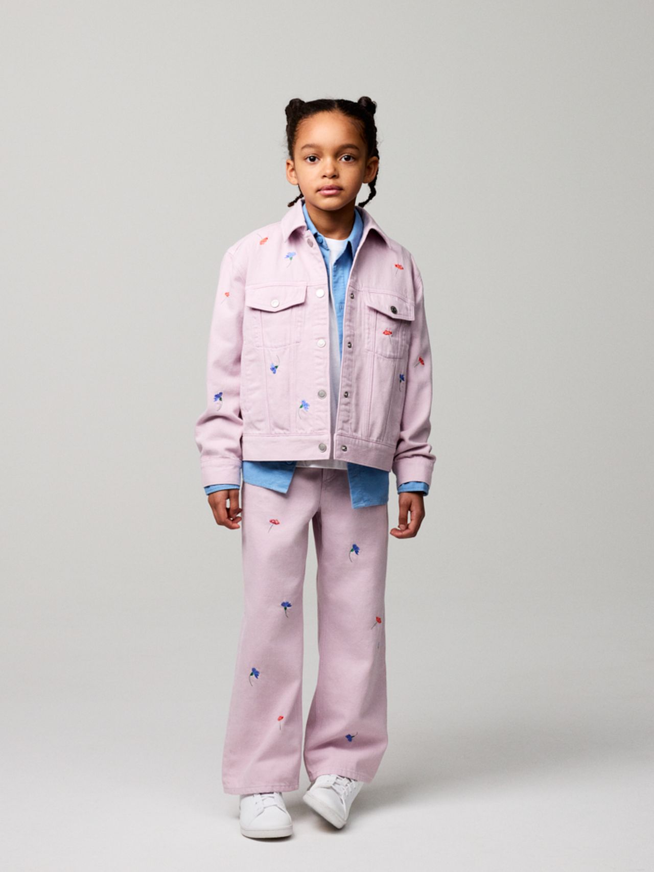 Child wearing a light pink jacket and pants with small embroidered designs, layered over a blue shirt, and white sneakers against a plain background.