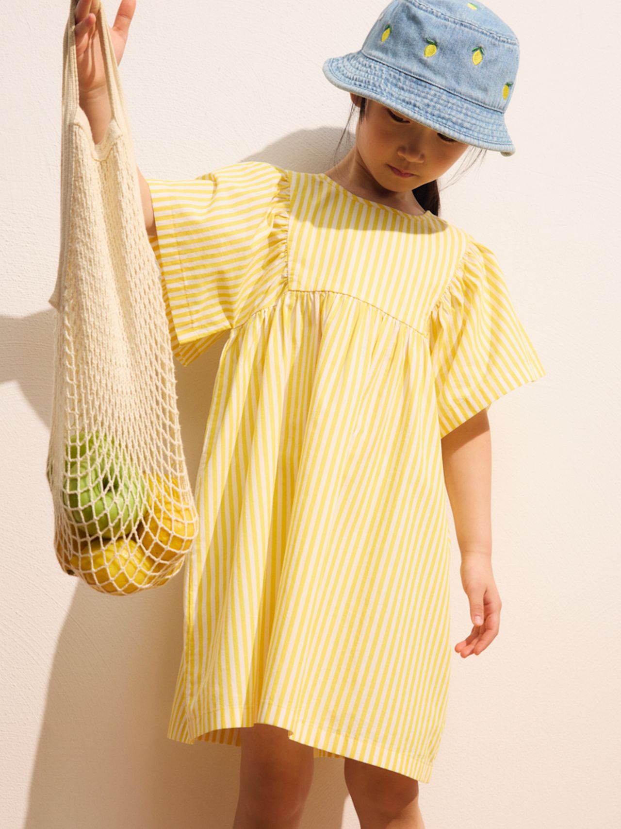 Child in a yellow striped dress and a denim bucket hat holds a mesh bag with fruit.