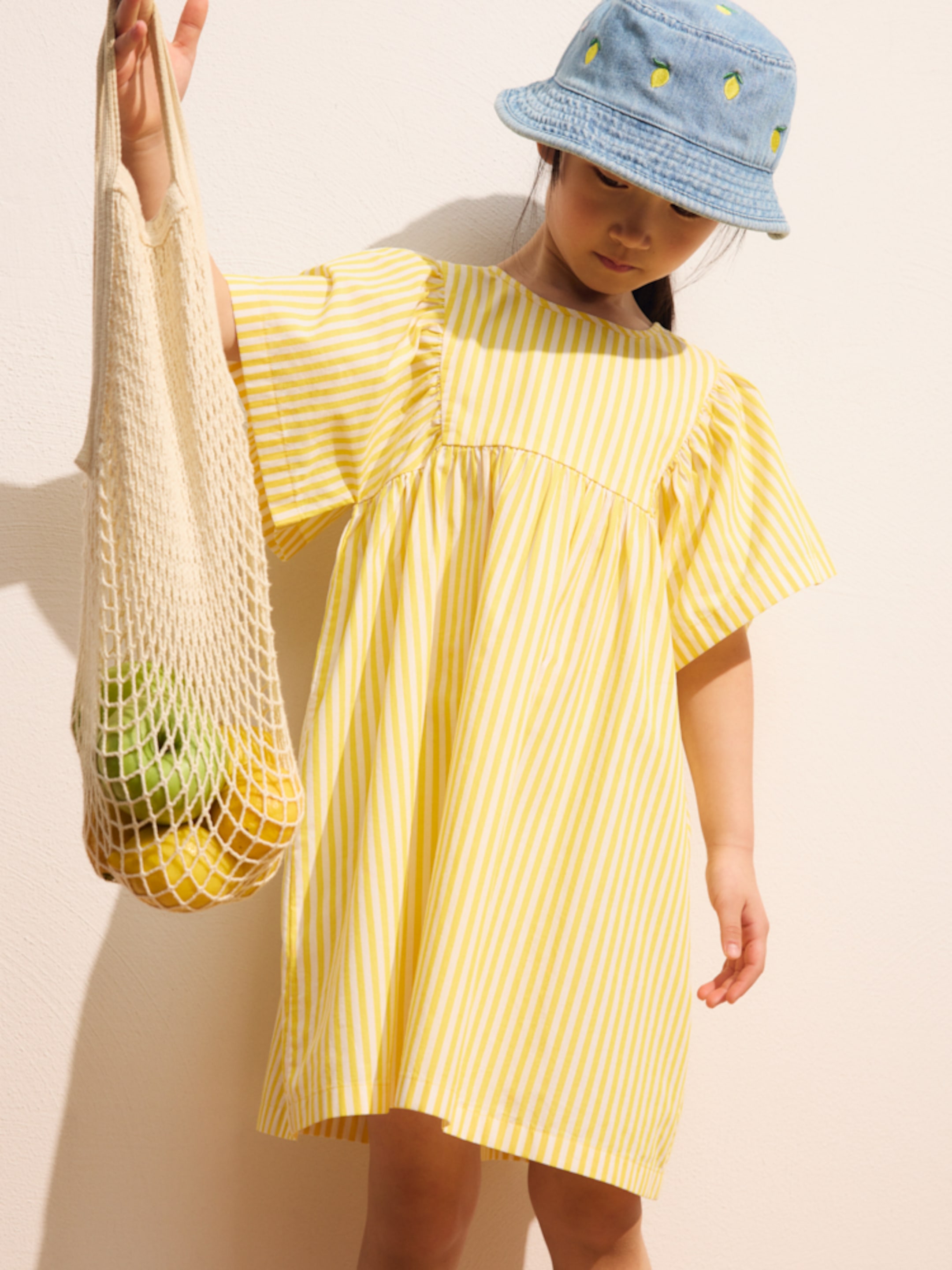 Child in a yellow striped dress and a denim bucket hat holds a mesh bag with fruit.
