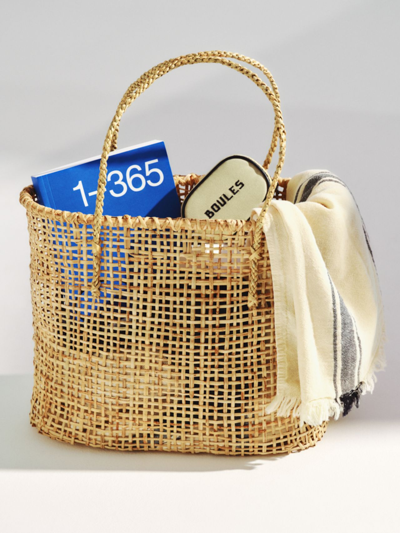 Woven basket containing a blue 1-365 box, "Boules" case, and a cream-striped fabric draped over the side.