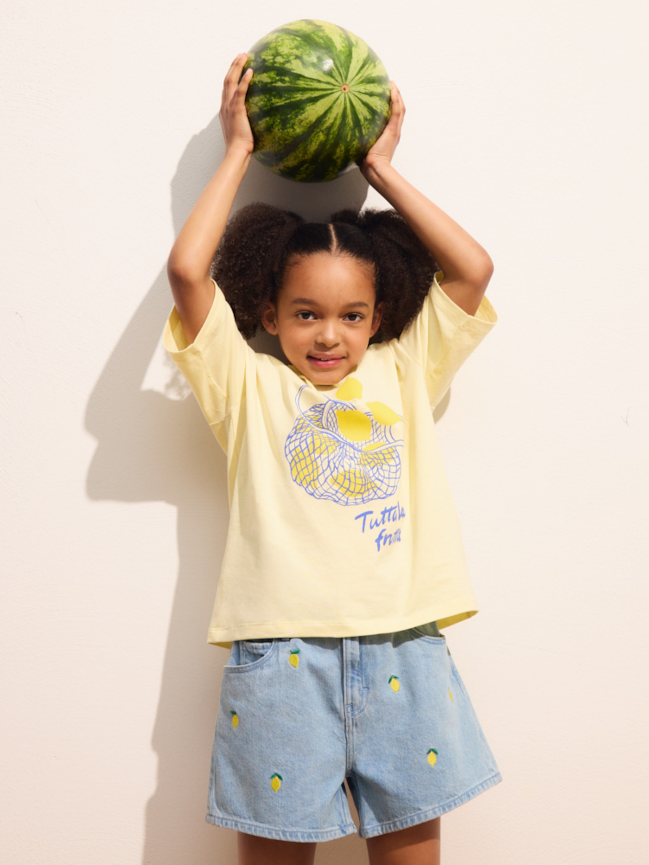 Child in a yellow shirt and denim shorts holds a watermelon over their head, smiling against a plain background.