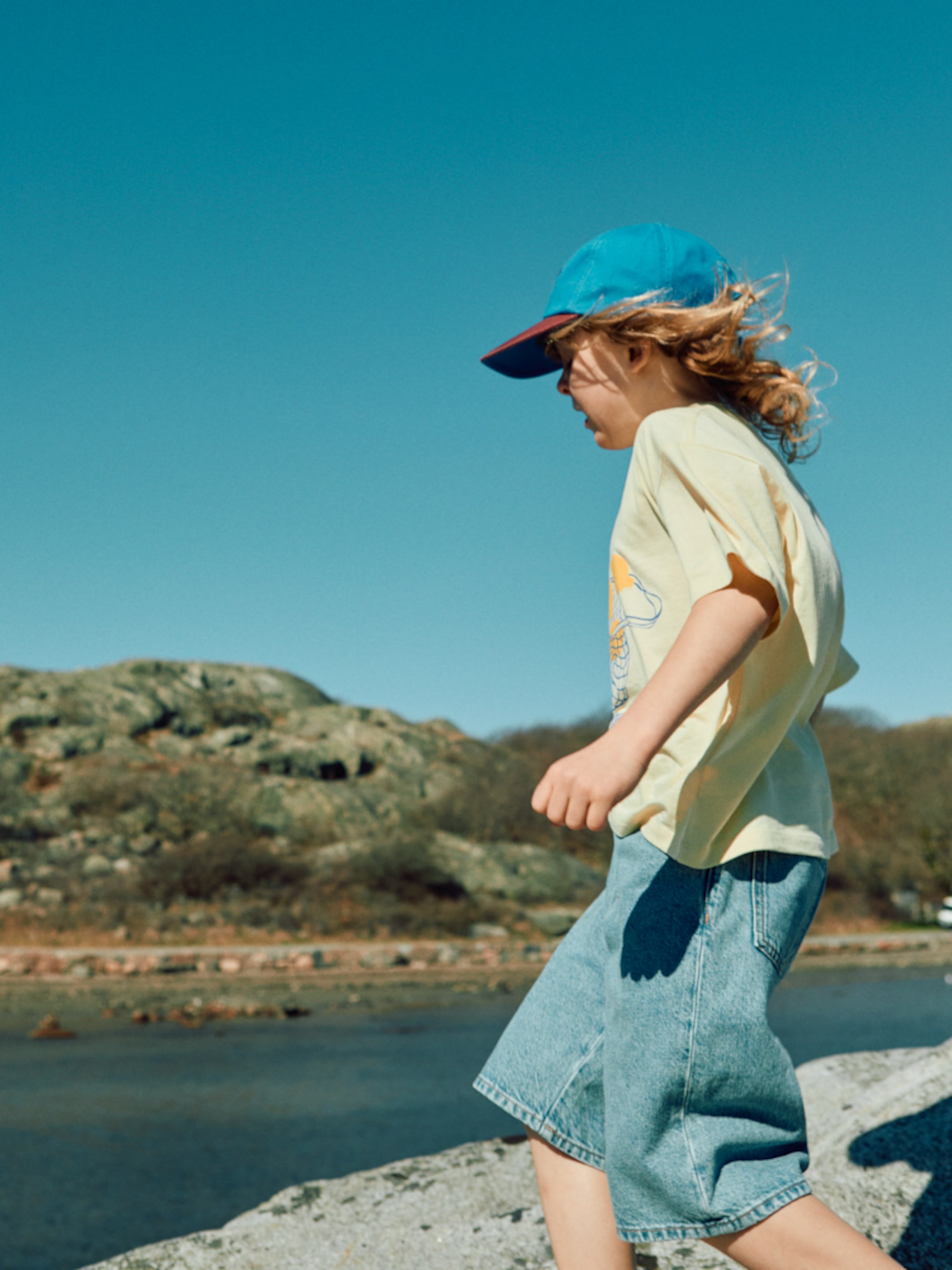 A child in a blue cap and yellow shirt walks on rocks by a river under a clear blue sky.