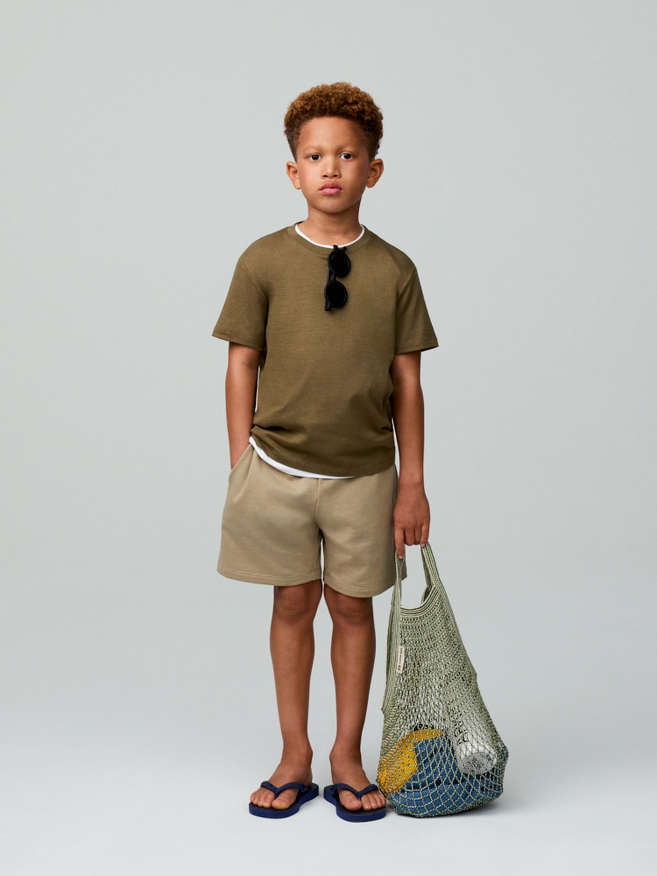 Young boy in a green shirt and beige shorts, wearing sunglasses, holds a mesh bag with a ball inside, standing against a plain background.