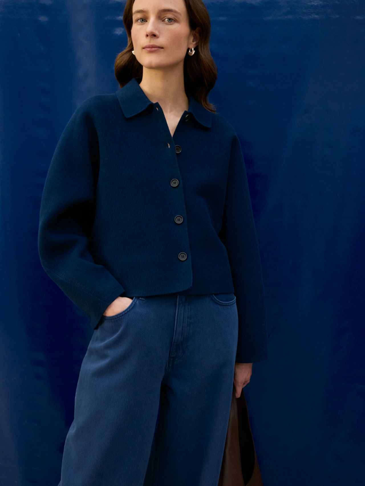 Woman in navy buttoned jacket and wide-legged jeans poses with one hand in pocket against a deep blue background.