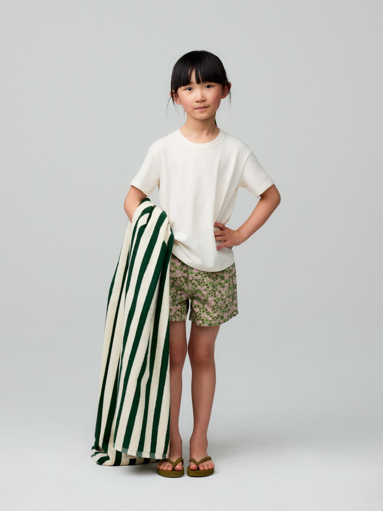 A young child in a white T-shirt and floral shorts holds a green-and-white striped towel, standing against a plain background.